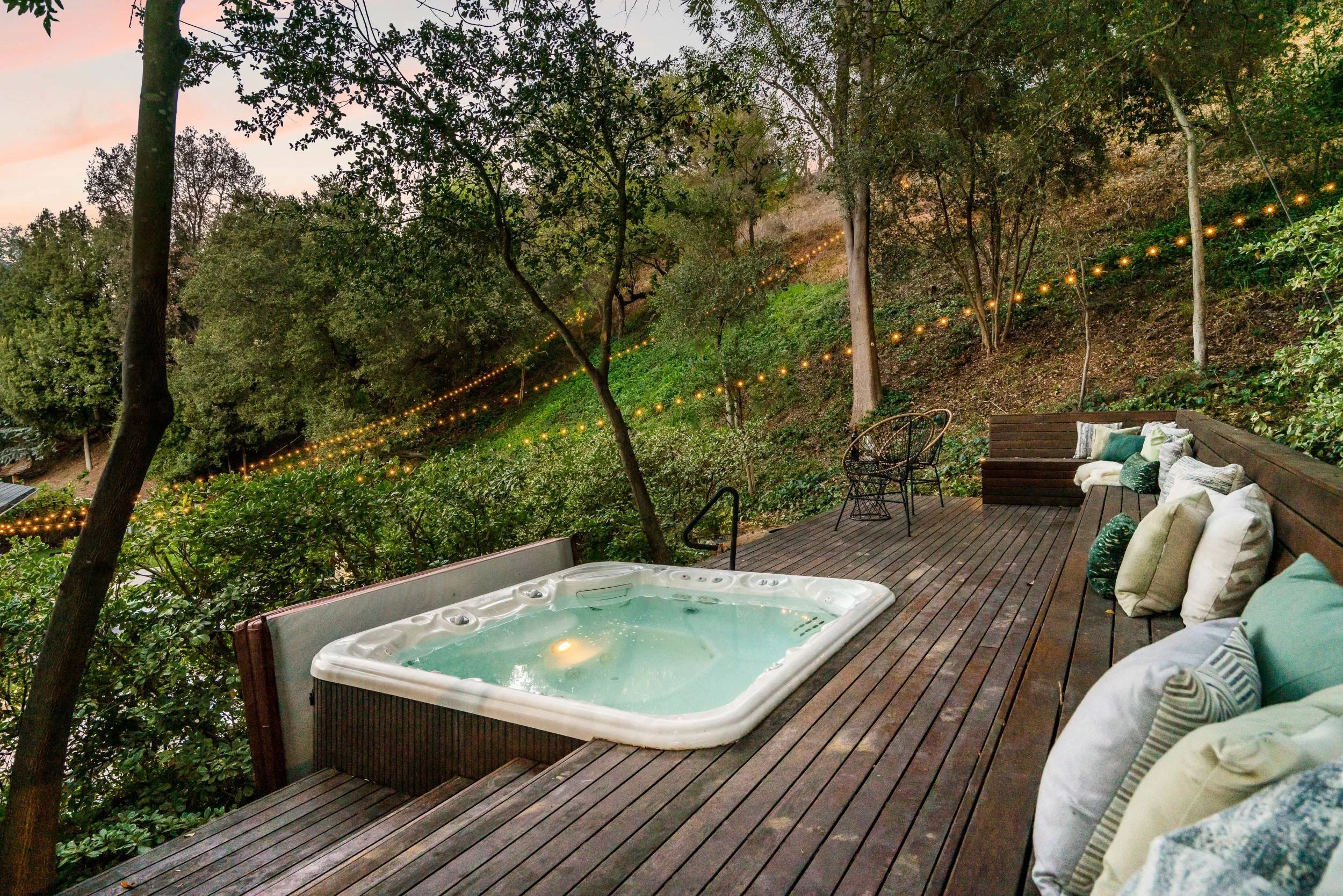 Outdoor deck with hot tub, seating area, and string lights overlooking a lush hillside covered in trees at dusk.