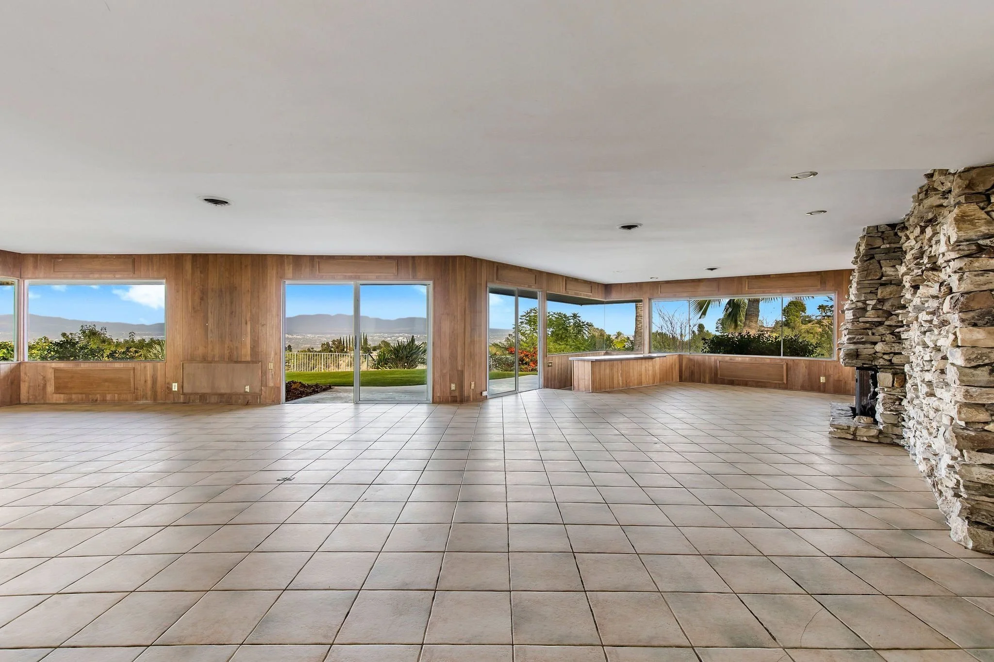 Spacious living room with large windows showing outdoor landscape, tiled floor, wooden wall paneling, stone fireplace, and sliding glass door opening to the garden.