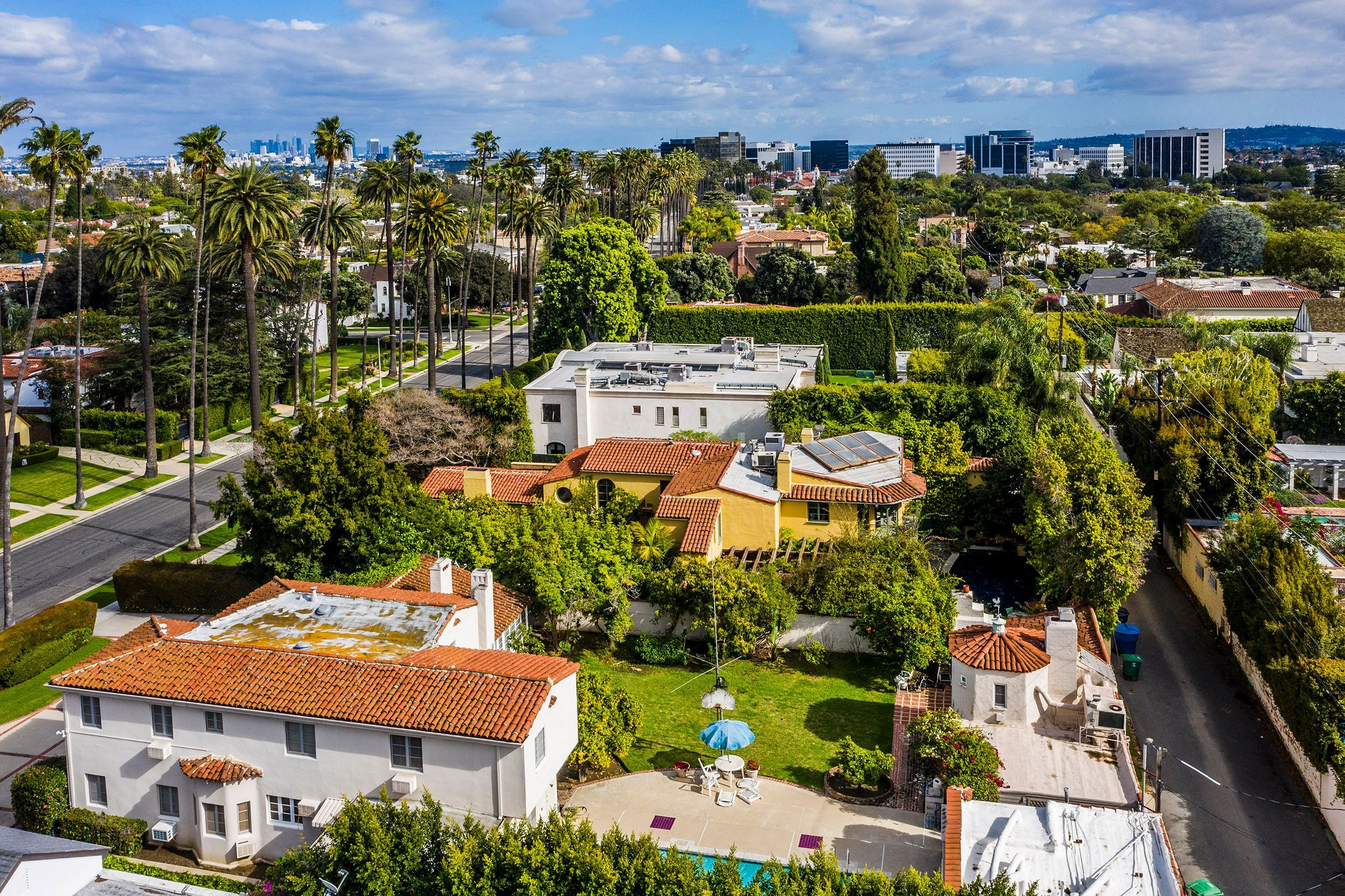 Aerial view of a residential area with lush greenery, tall palm trees, colorful houses with tile roofs, and high-rise buildings in the background under a partly cloudy sky.
