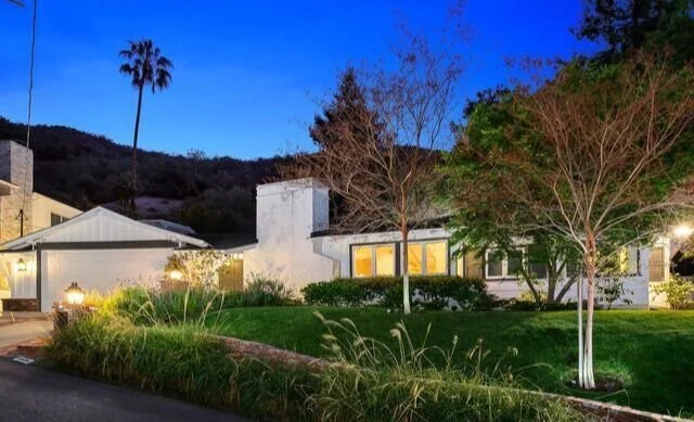 Exterior view of a house during twilight with illuminated windows, surrounded by trees and landscaping, with a mountain and blue sky in the background.