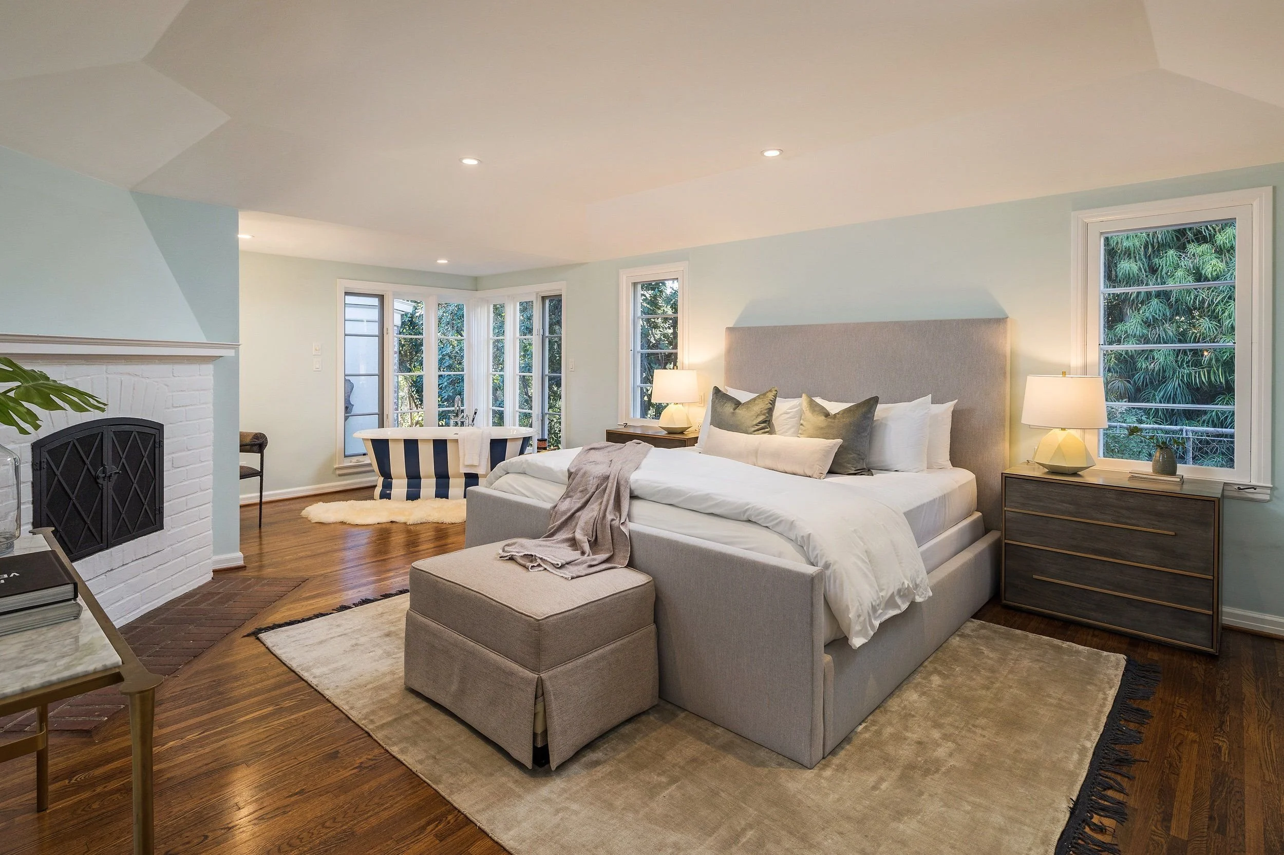 A cozy and modern bedroom with a large bed, multiple pillows, bedside tables with lamps, a beige ottoman, a striped armchair, large windows with greenery outside, a white brick fireplace, and hardwood floors.