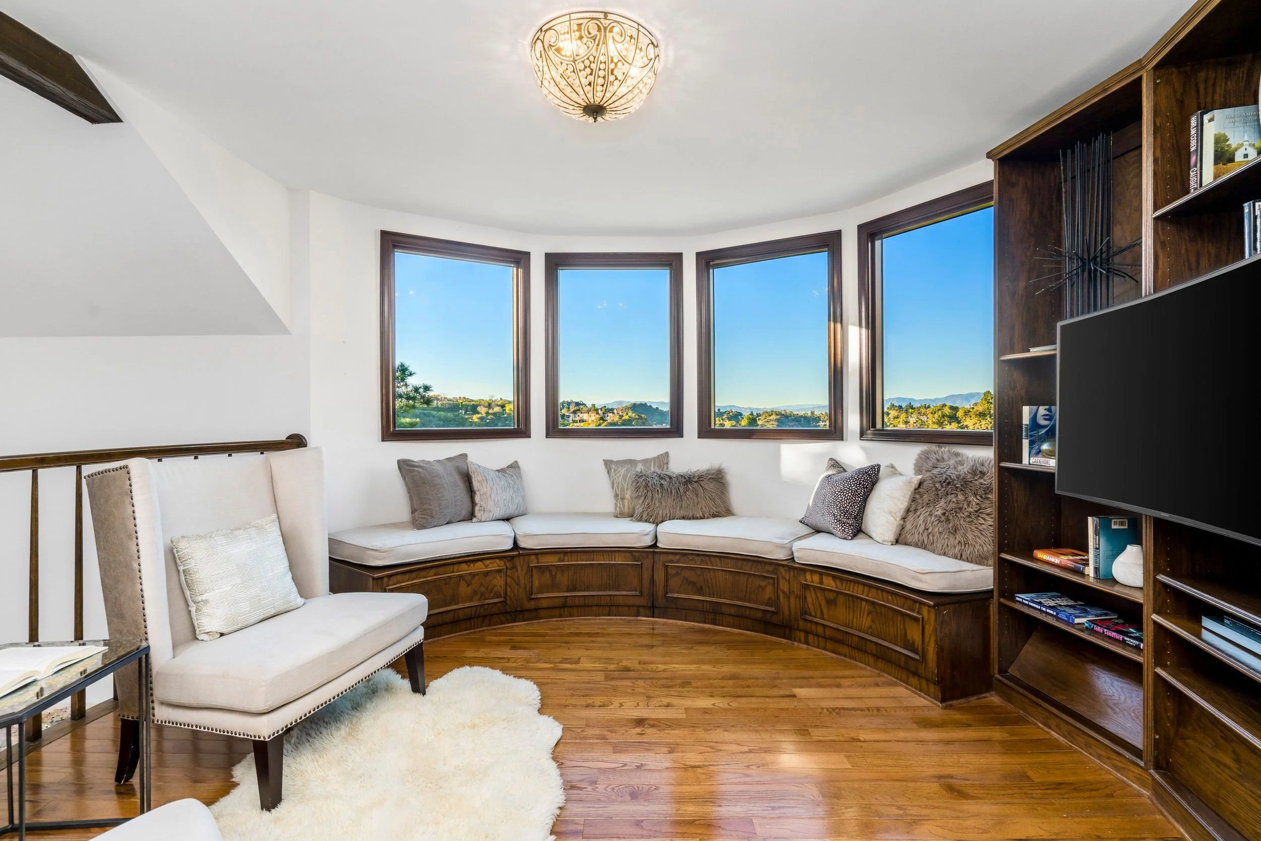 A cozy living room with a large white sectional sofa decorated with various pillows, a white armchair, and a fluffy rug. There is a built-in wooden bookcase filled with books and decor, large bay windows showing a scenic landscape outside, and a deco
