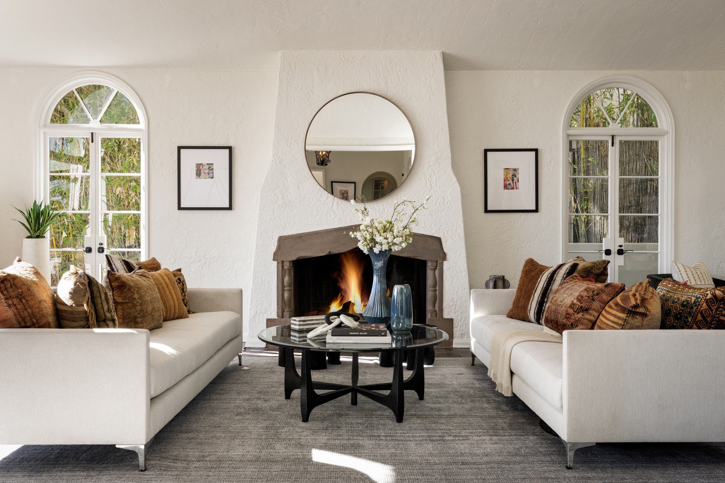 Living room with a fireplace, two white sofas with multicolored cushions, a round coffee table with decorative vases and books, framed artwork on the wall, a round mirror above the fireplace, and large arched windows with greenery outside.