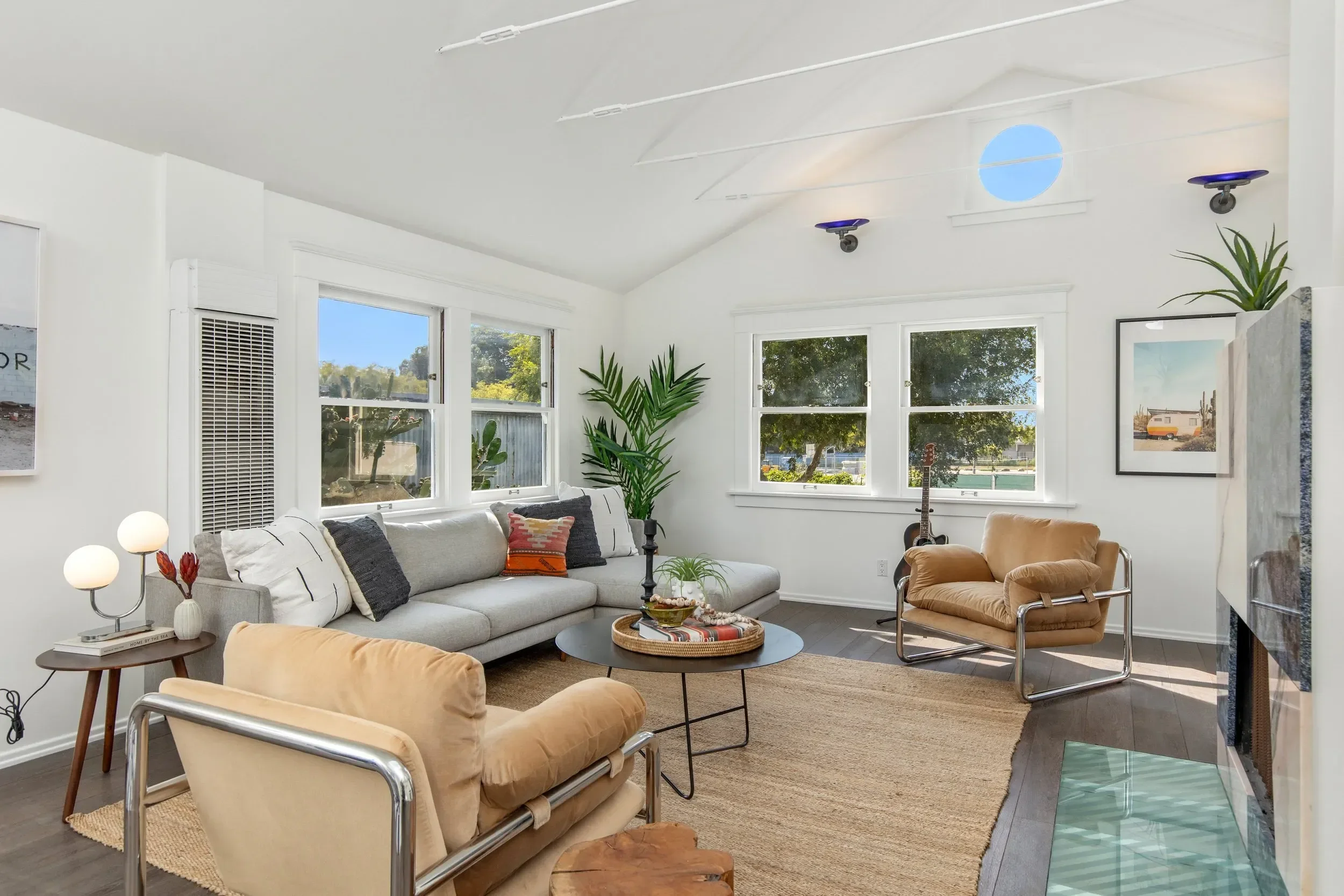 A bright living room with a white sectional sofa, throw pillows, potted plants, and a guitar by the window. Large windows let in natural light, and there are two framed pictures on the walls. A beige armchair is nearby, and there is a round coffee ta