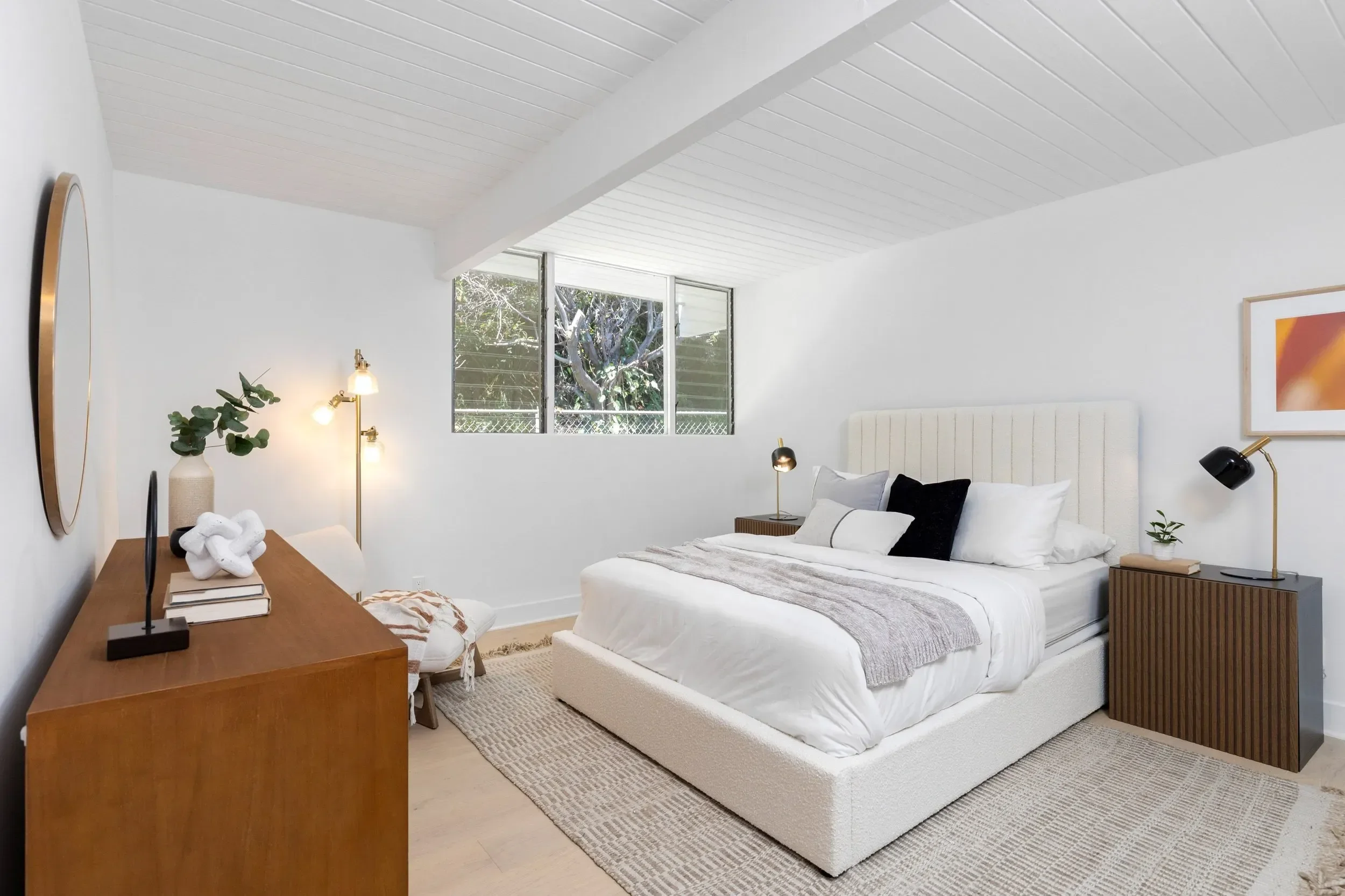 A bedroom with a white bed, wooden nightstand, abstract artwork on the wall, window with trees outside, beige rug, and various lamps and decor items.
