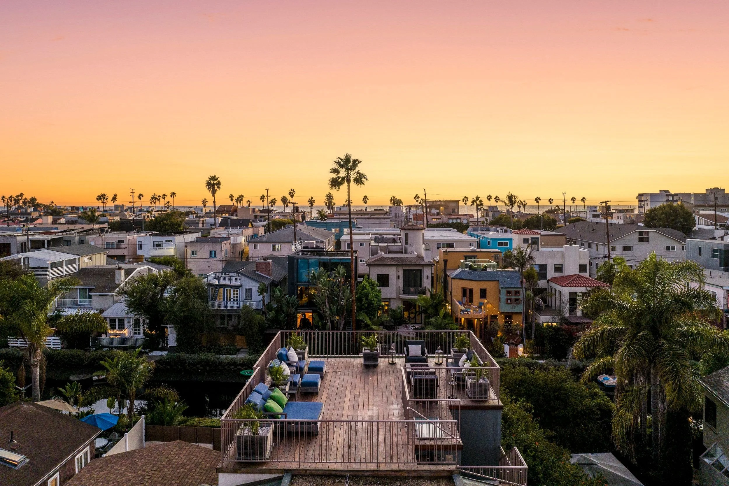 View of a residential neighborhood at sunset with houses, palm trees, and a clear sky in the background. In the foreground, a wooden deck with outdoor furniture and lounge chairs.