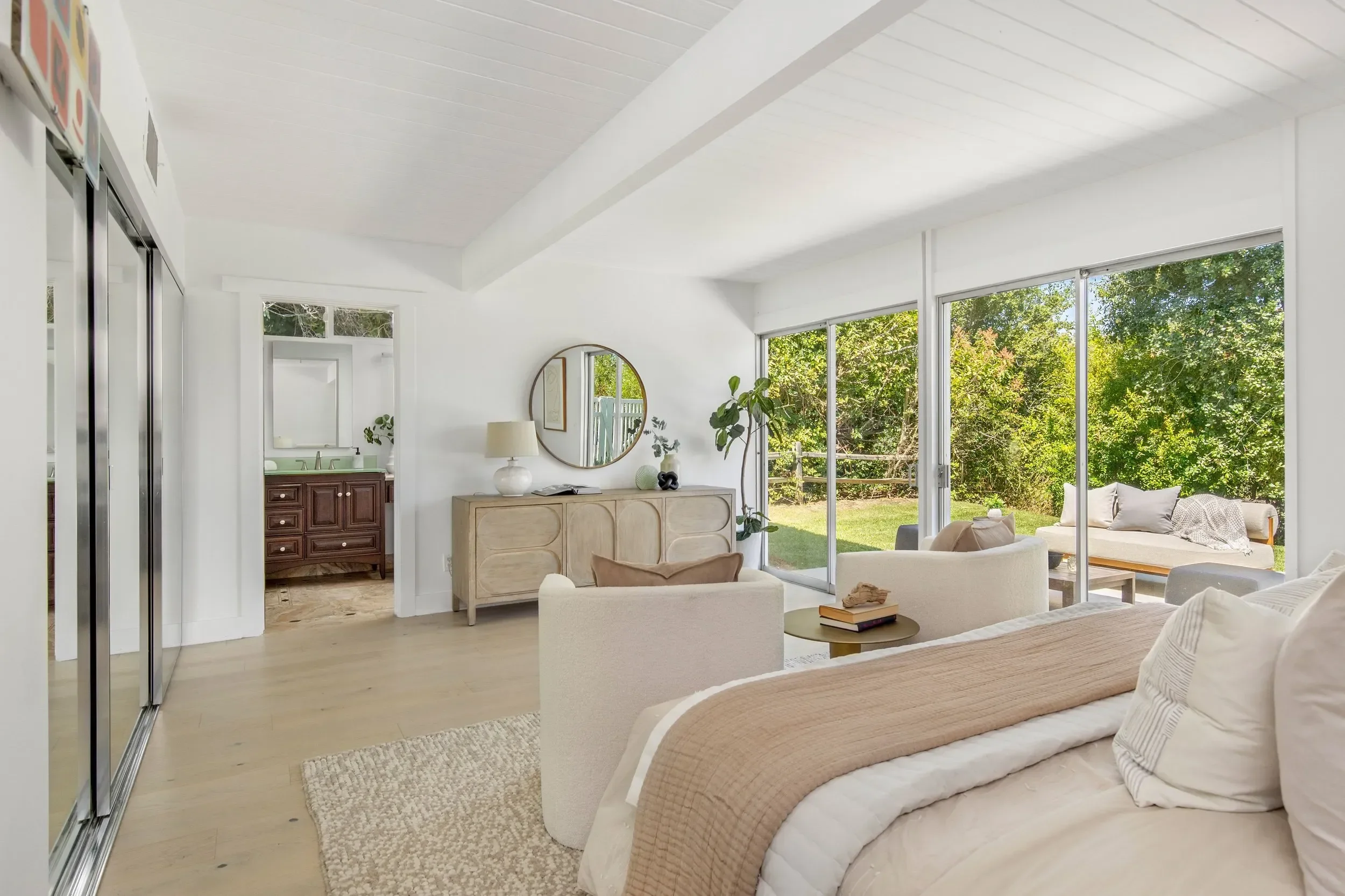 Living room with large sliding glass doors revealing a green backyard, white walls, ceiling, and furniture including a beige couch with cushions, a round coffee table with books, a wooden sideboard with decorative items, and a mirror on the wall.