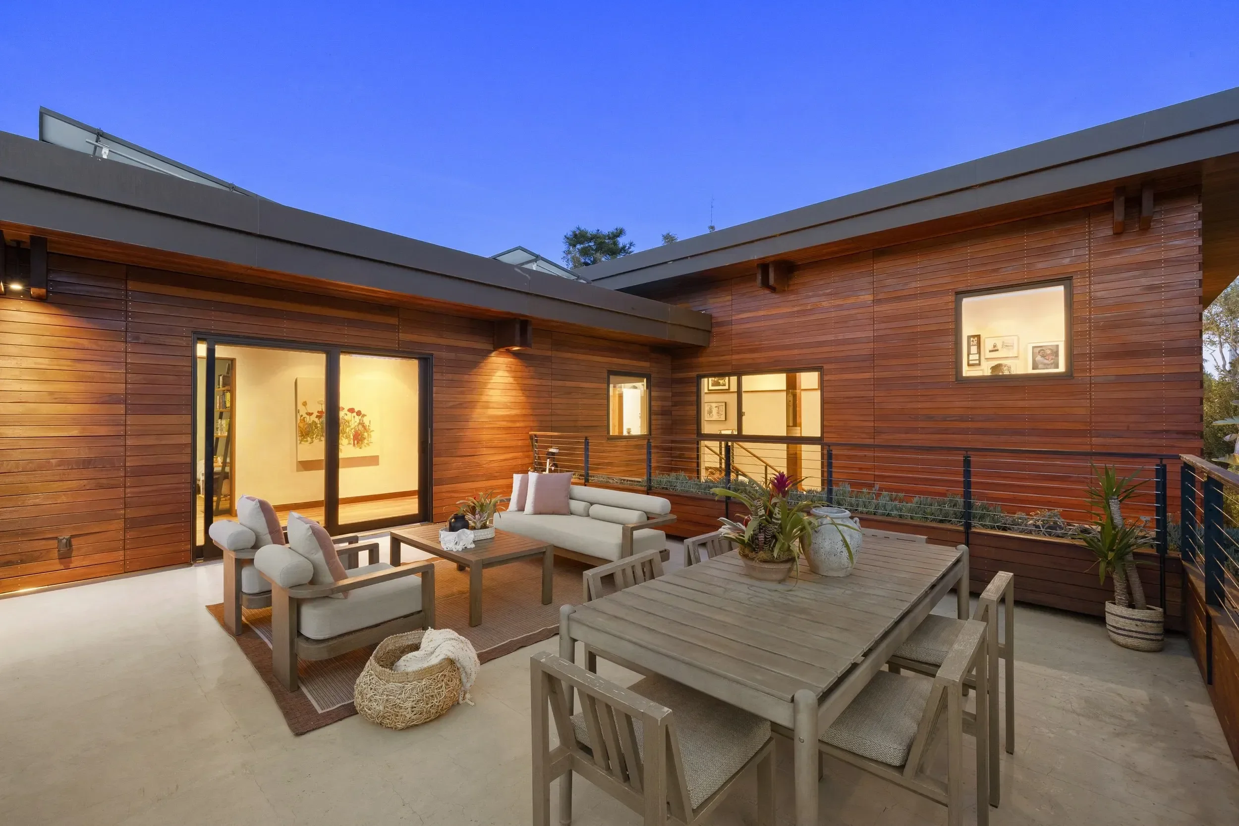 Exterior view of a modern house with a rooftop deck furnished with outdoor seating and potted plants, during evening with a clear blue sky.