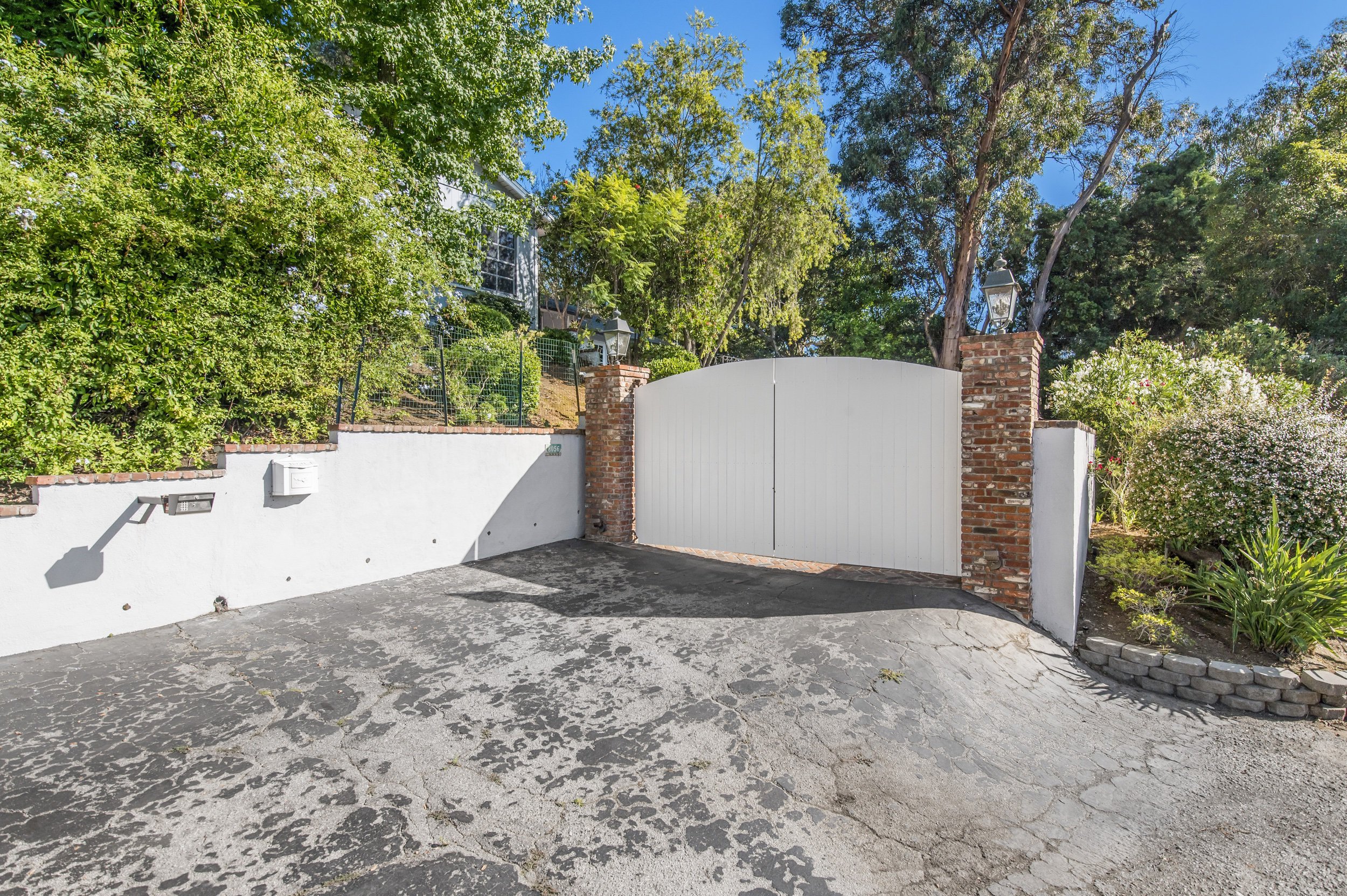 A white gate with brick pillars on each side and two lanterns on top of the pillars. The gate is located in front of a house behind a white wall, with a driveway in the front. There is greenery and trees surrounding the area.