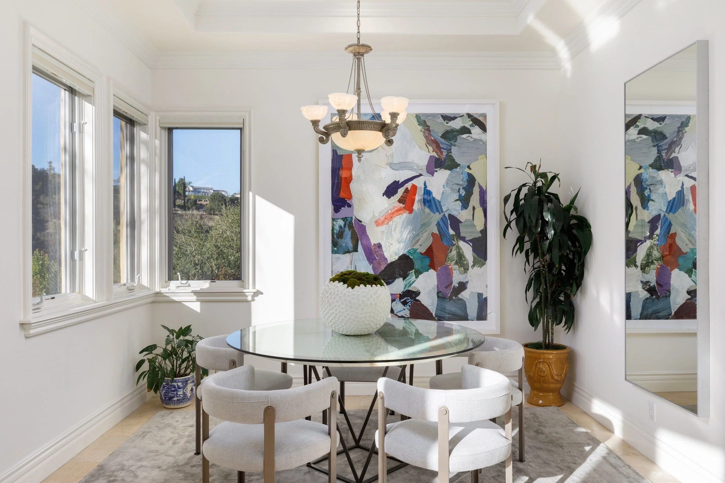 A bright dining room with white walls and large windows. A round glass table with white upholstered chairs surrounds a white vase with moss. There are abstract colorful paintings on the walls, and a potted plant in a yellow pot beside the paintings. 