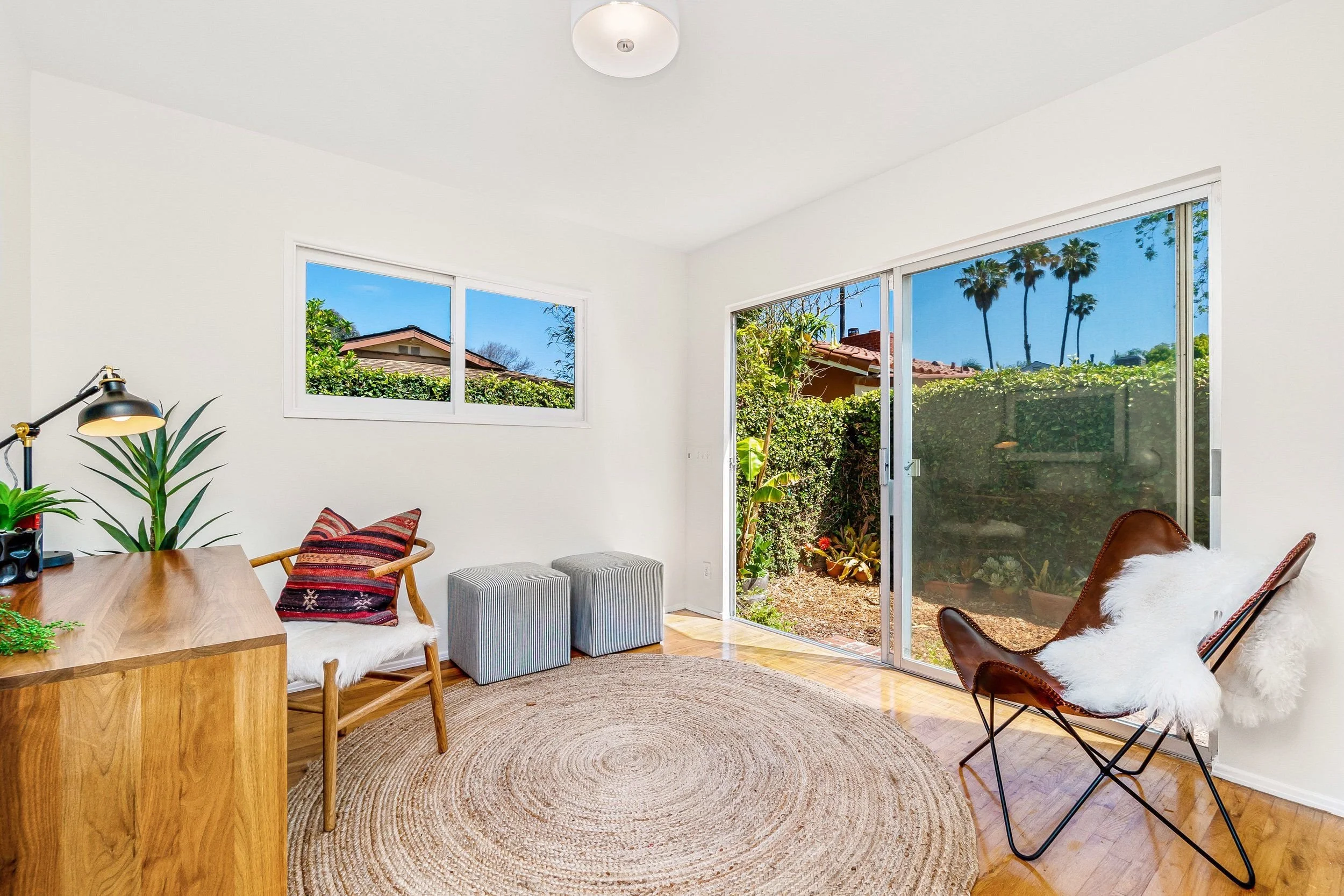 Bright living room with large sliding glass door and window showing a backyard with plants and tall trees, including palm trees.