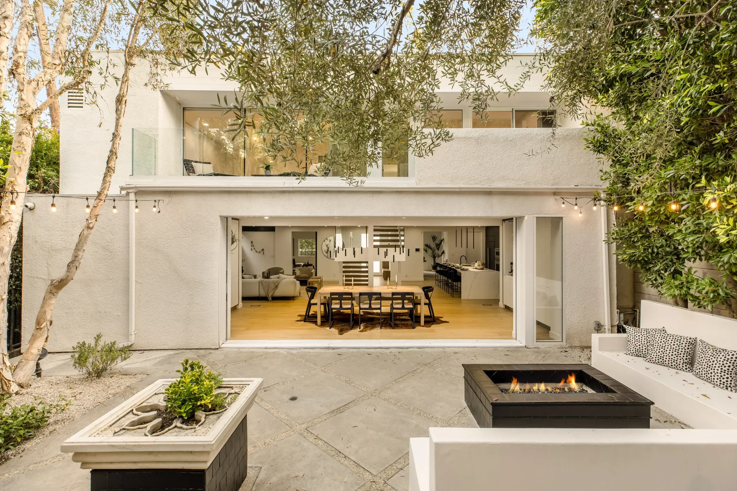 View of a modern two-story house with large glass doors, an outdoor patio, and string lights, featuring a garden with plants and trees. Inside, an open-concept living and dining area with wooden flooring and contemporary furniture is visible through 