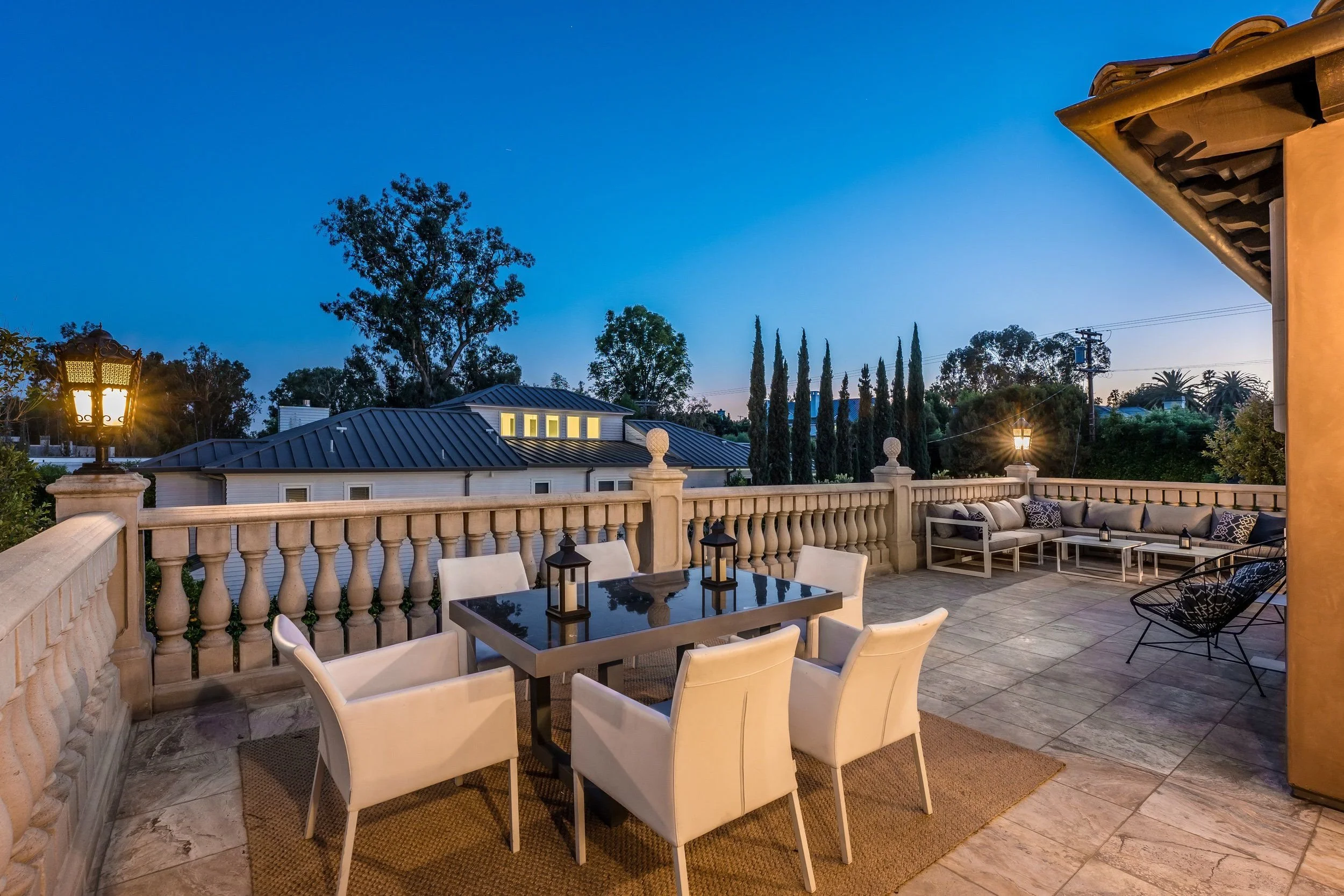 An outdoor balcony or terrace with a dining table, chairs, and a sitting area, overlooking neighboring houses, trees, and a clear evening sky with streetlights turned on.