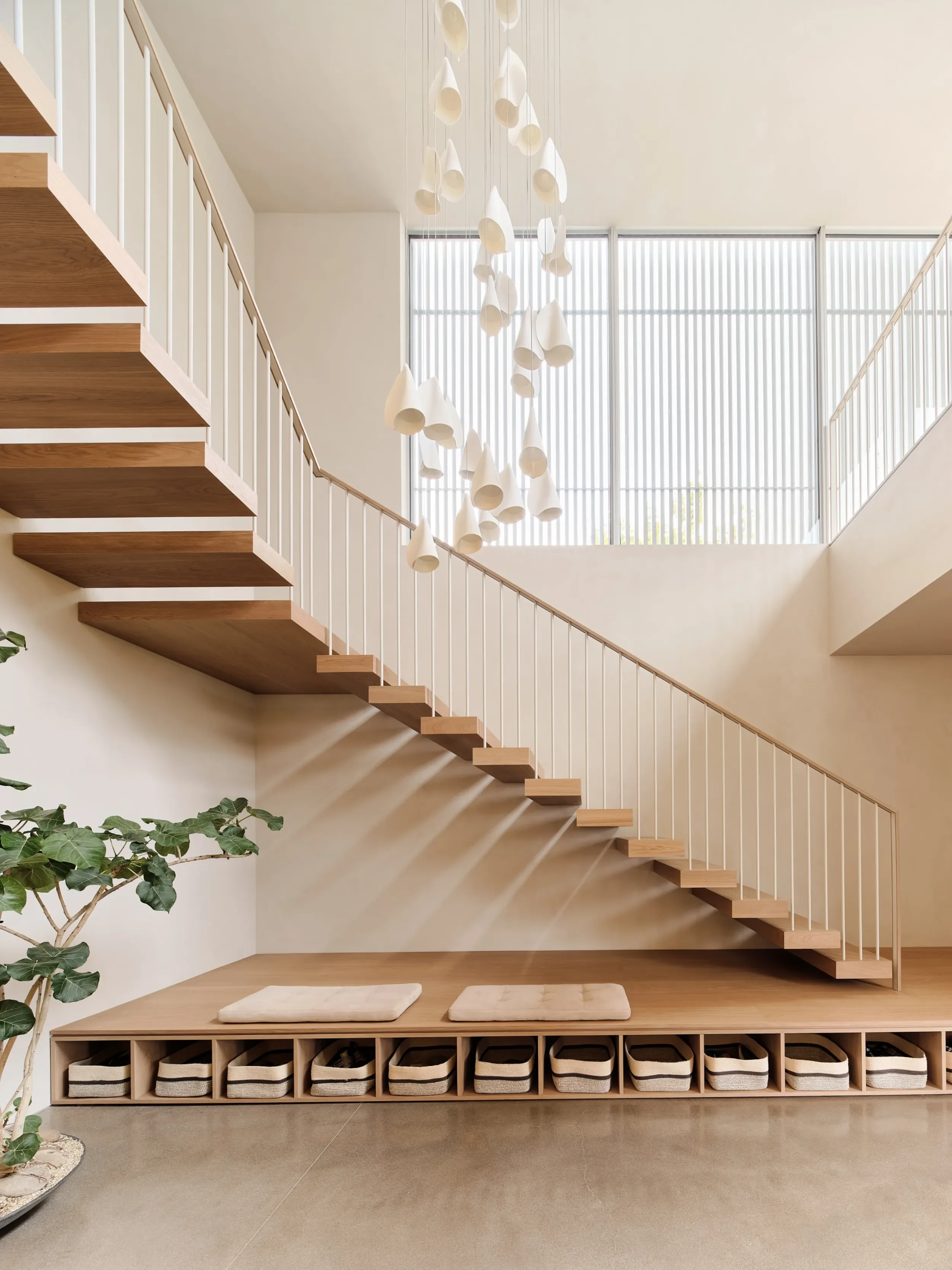 Modern staircase with wooden steps and white railing in a bright, airy interior. A contemporary chandelier with white, bell-shaped fixtures hangs from the ceiling, and large windows with vertical blinds allow natural light to fill the space. A wooden