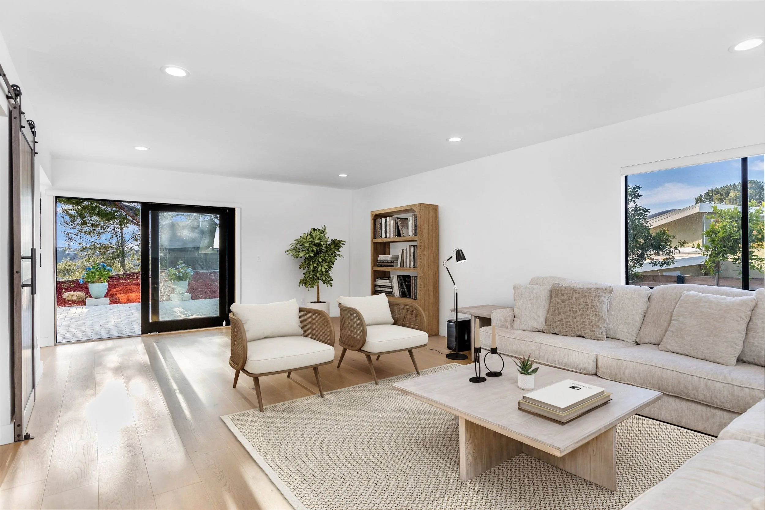 Living room with light wood flooring, a beige sectional sofa, two cream armchairs, a wooden bookshelf, a black floor lamp, a coffee table with books and a small plant, large windows showing trees outside, and a sliding glass door leading to a patio w