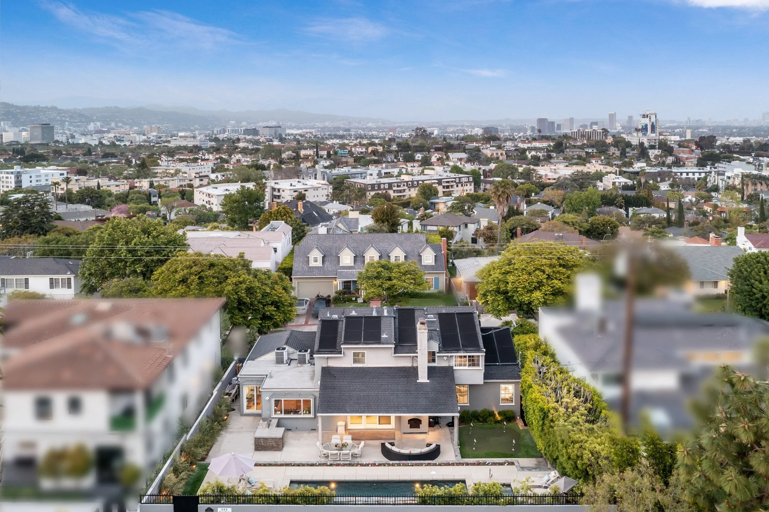 Aerial view of a residential neighborhood in a city with a skyline in the background, lush green trees, and clear blue sky.
