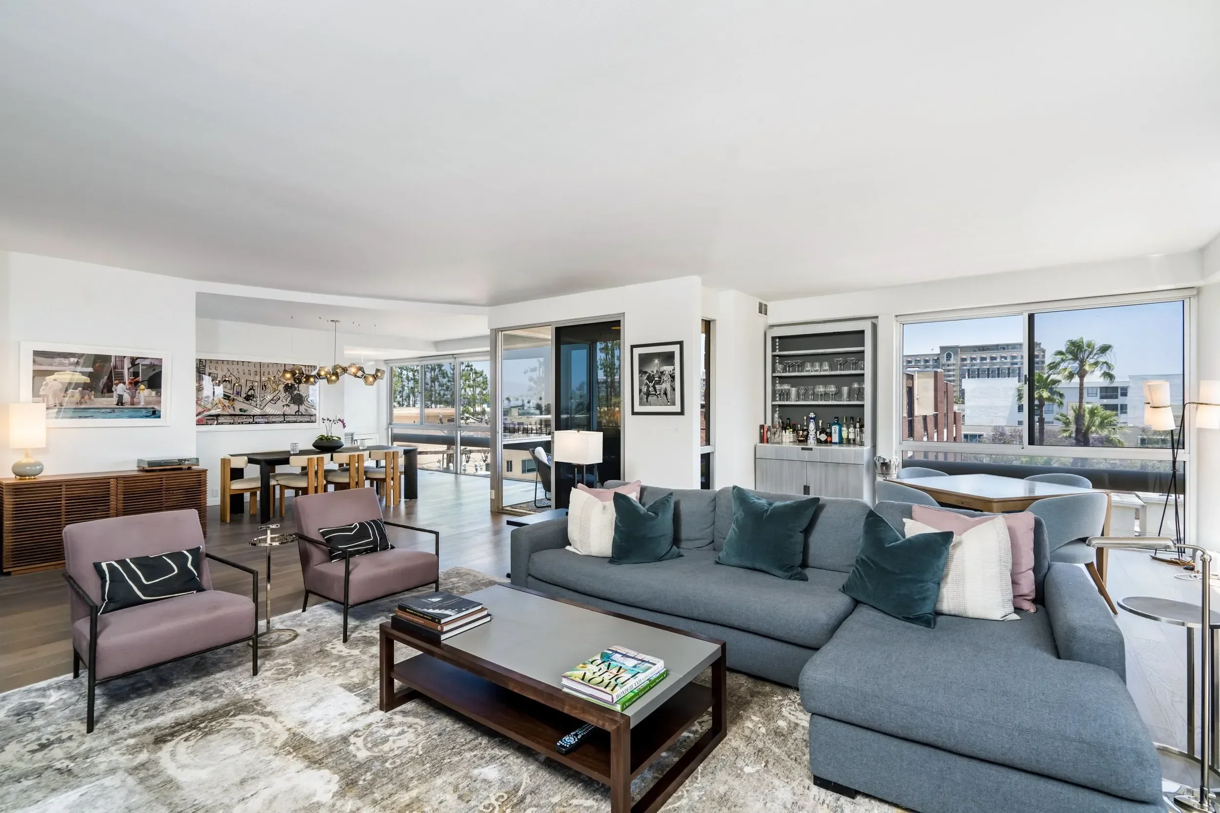 A modern, open-concept living room with large windows, a gray sofa, pink armchairs, a wooden coffee table with magazines, and a dining area with a dining table and chairs. The space is decorated with contemporary art and lighting fixtures.