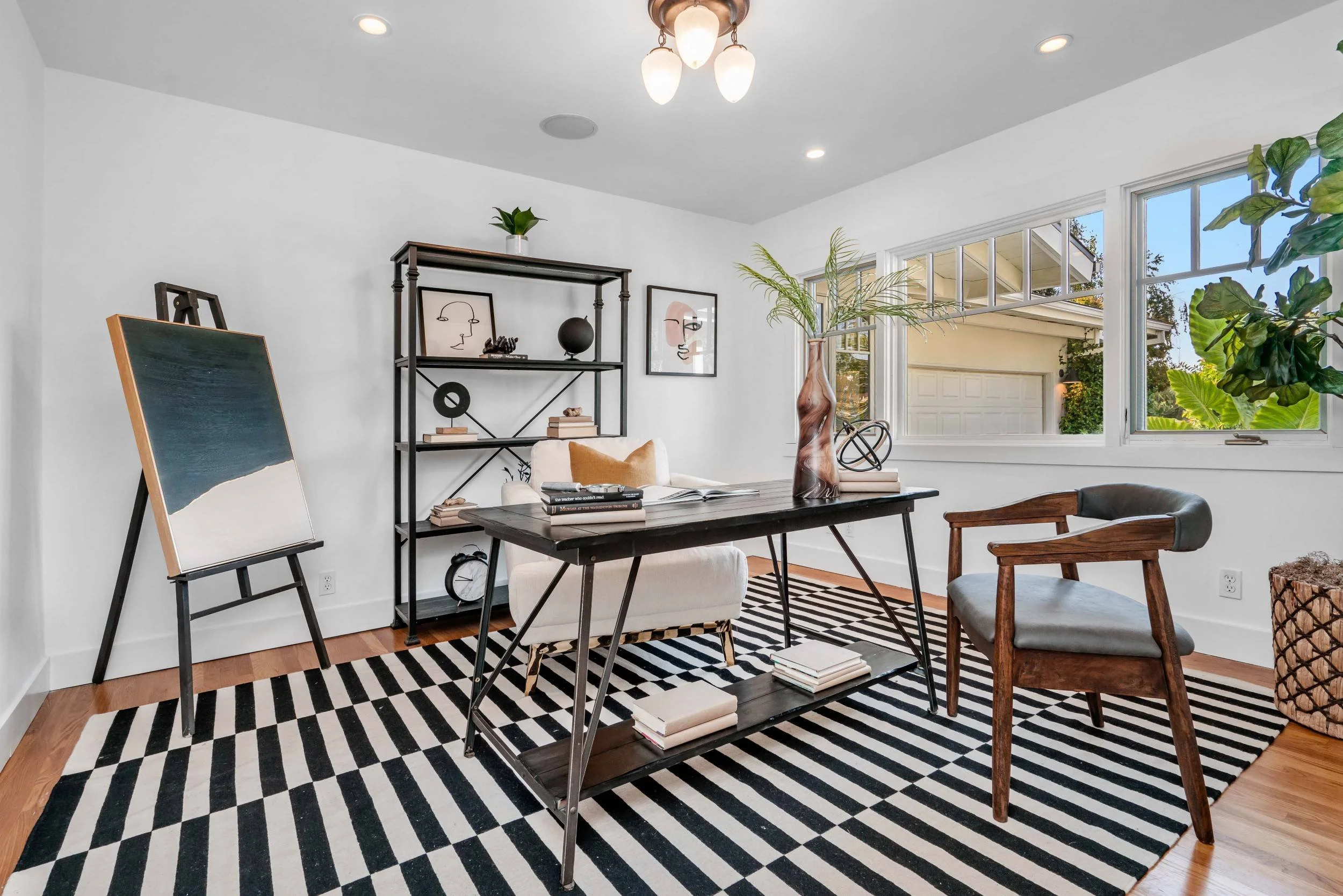 A bright modern living room with a patterned black and white striped rug, a black desk with books and a decorative vase, an easel with a painting, a white armchair, a black metal shelving unit with artwork and decor, large windows with a view of a ye
