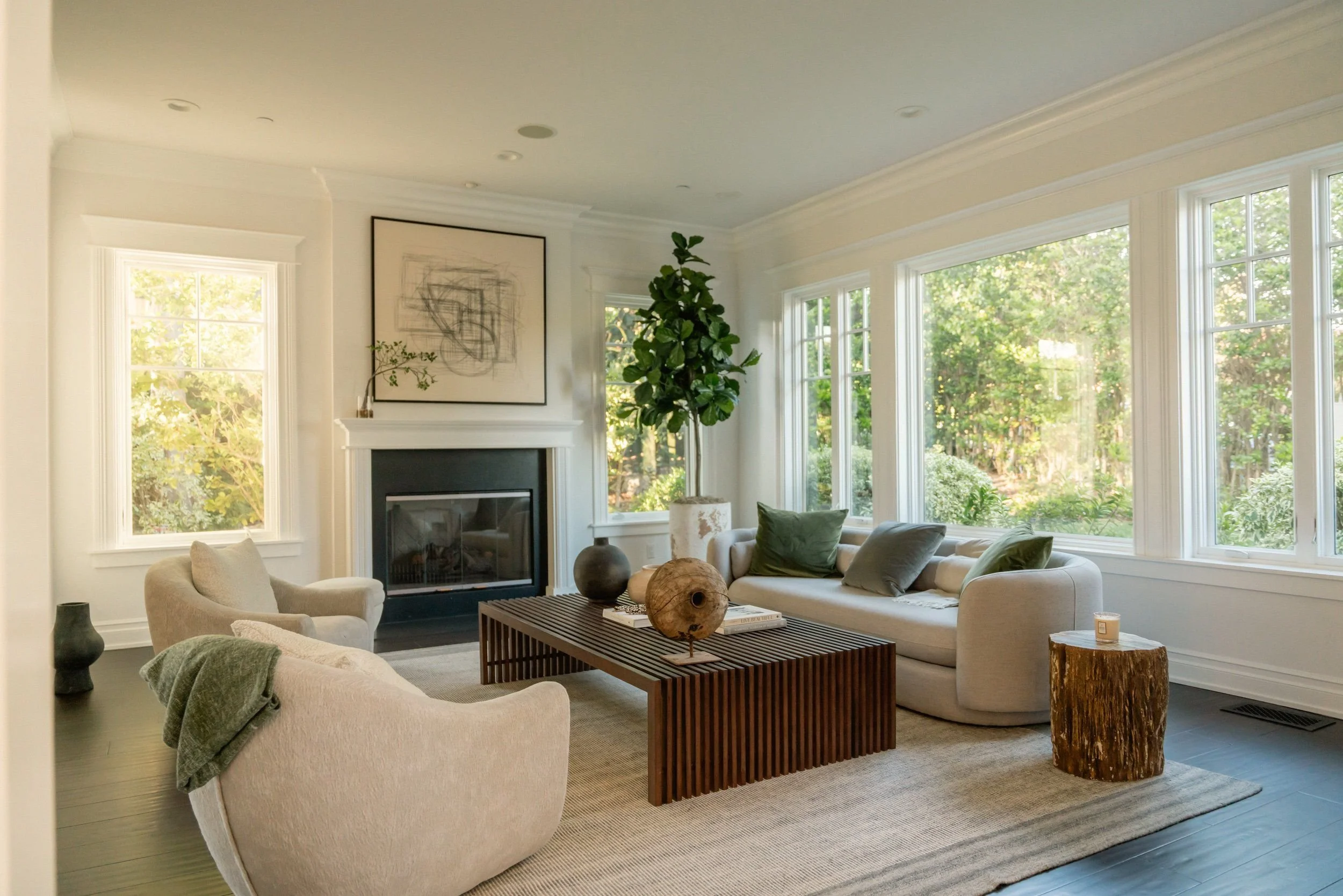 A bright living room with large windows showing green trees outside, a fireplace with a framed abstract artwork above, a tall leafy plant, beige sofas, a wooden coffee table with decorative objects, and a beige armchair with a green blanket.