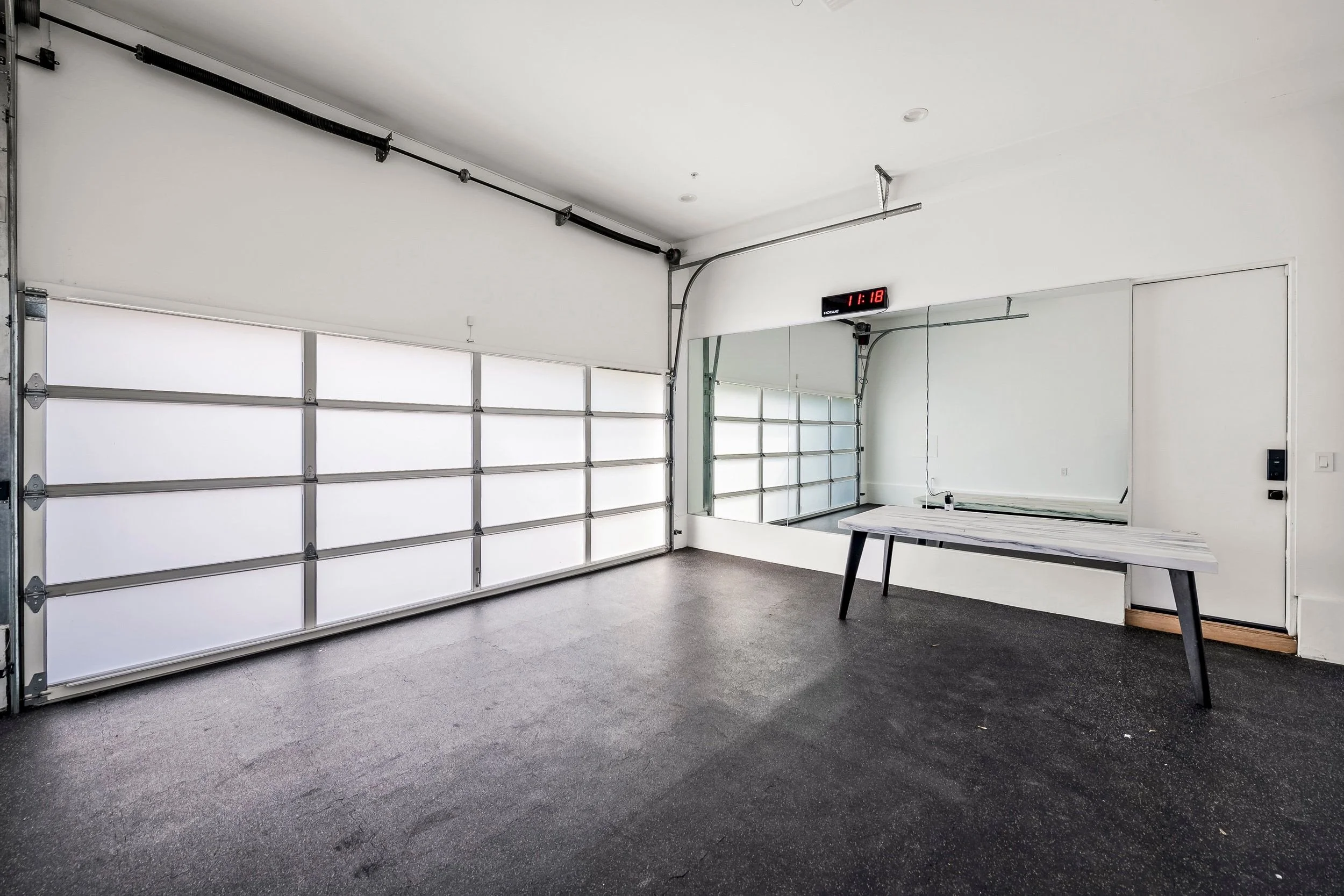 A clean, empty garage with white walls, a closed white garage door, and a black digital clock showing 11:18. There is a table with a marble top and black legs in the corner and a mirror on the wall reflecting part of the garage.