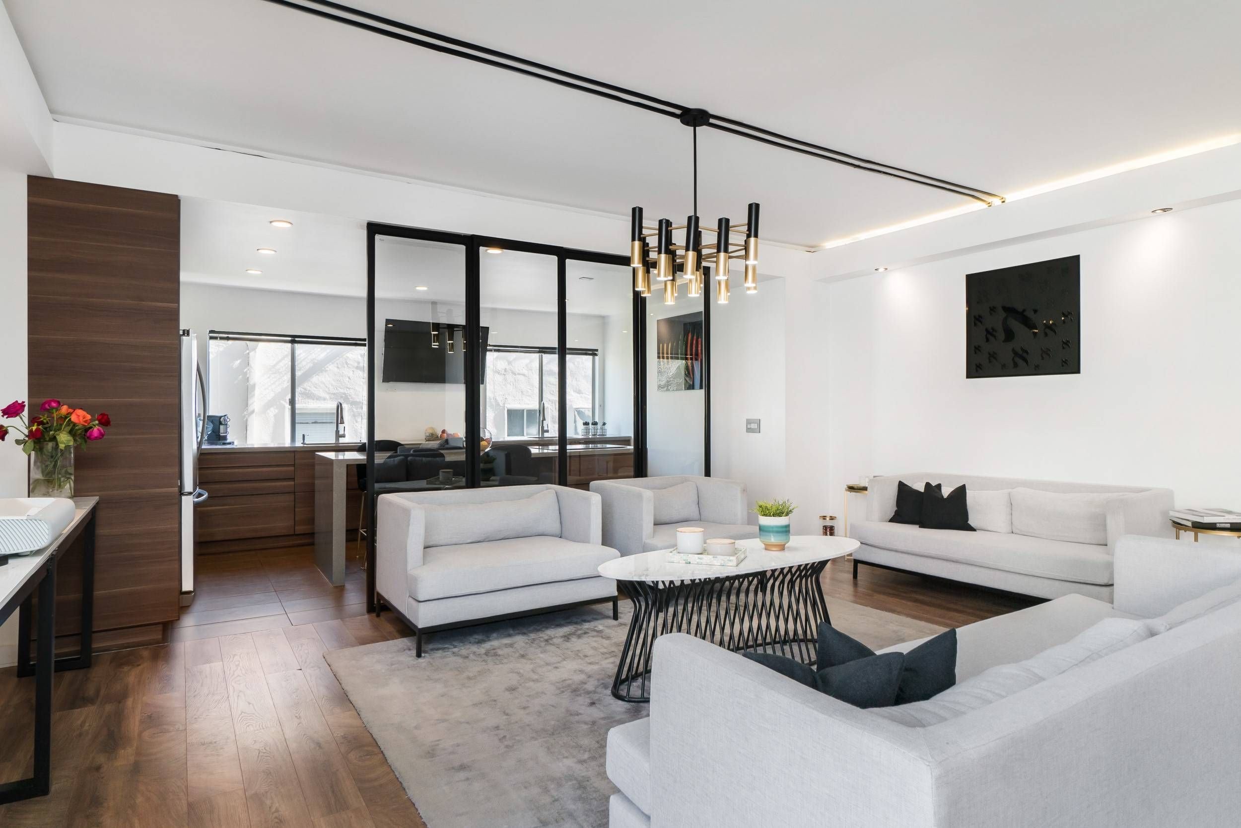 Modern living room with white sofas, a black metal coffee table, wooden flooring, and a chandelier with multiple gold and black lights, with a kitchen area in the background.