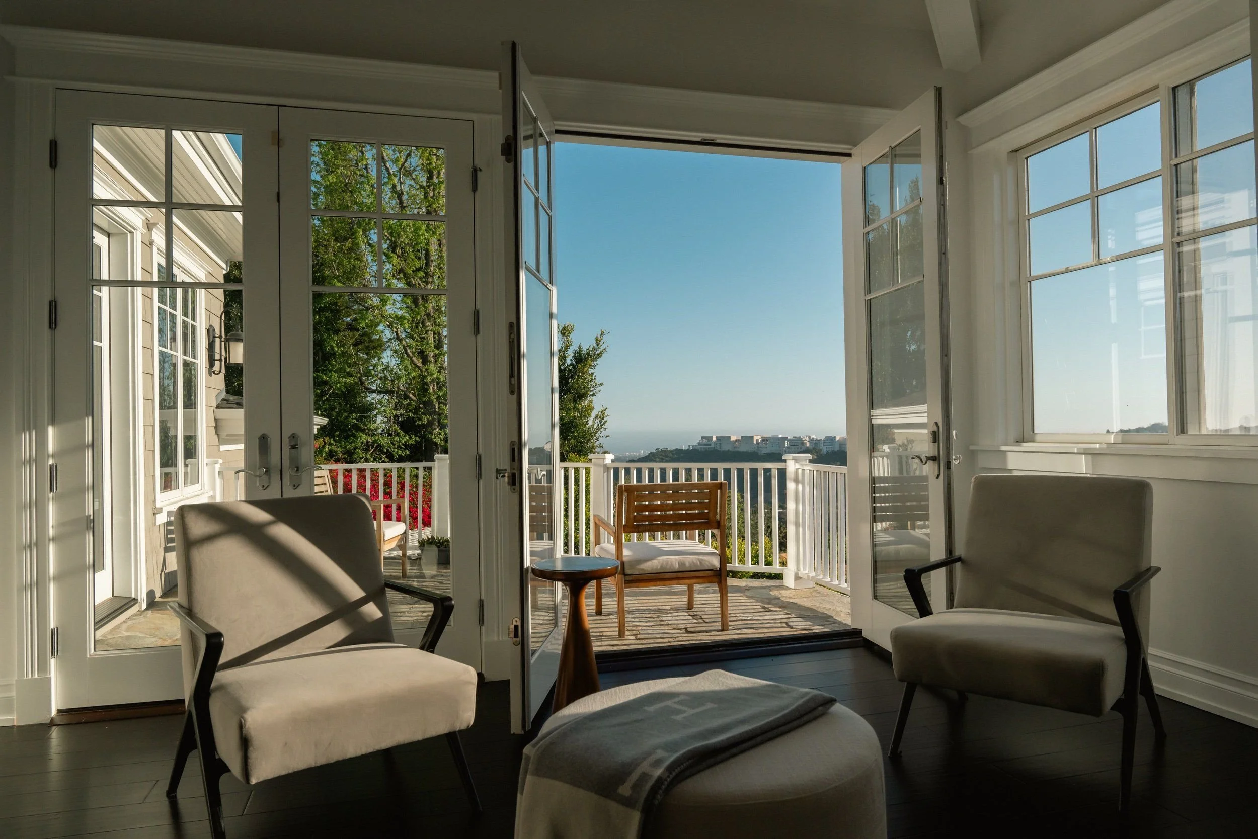 Sunlit porch with white French doors open to a view of green trees, a blue sky, and distant white buildings, furnished with white chairs and a small wooden table.