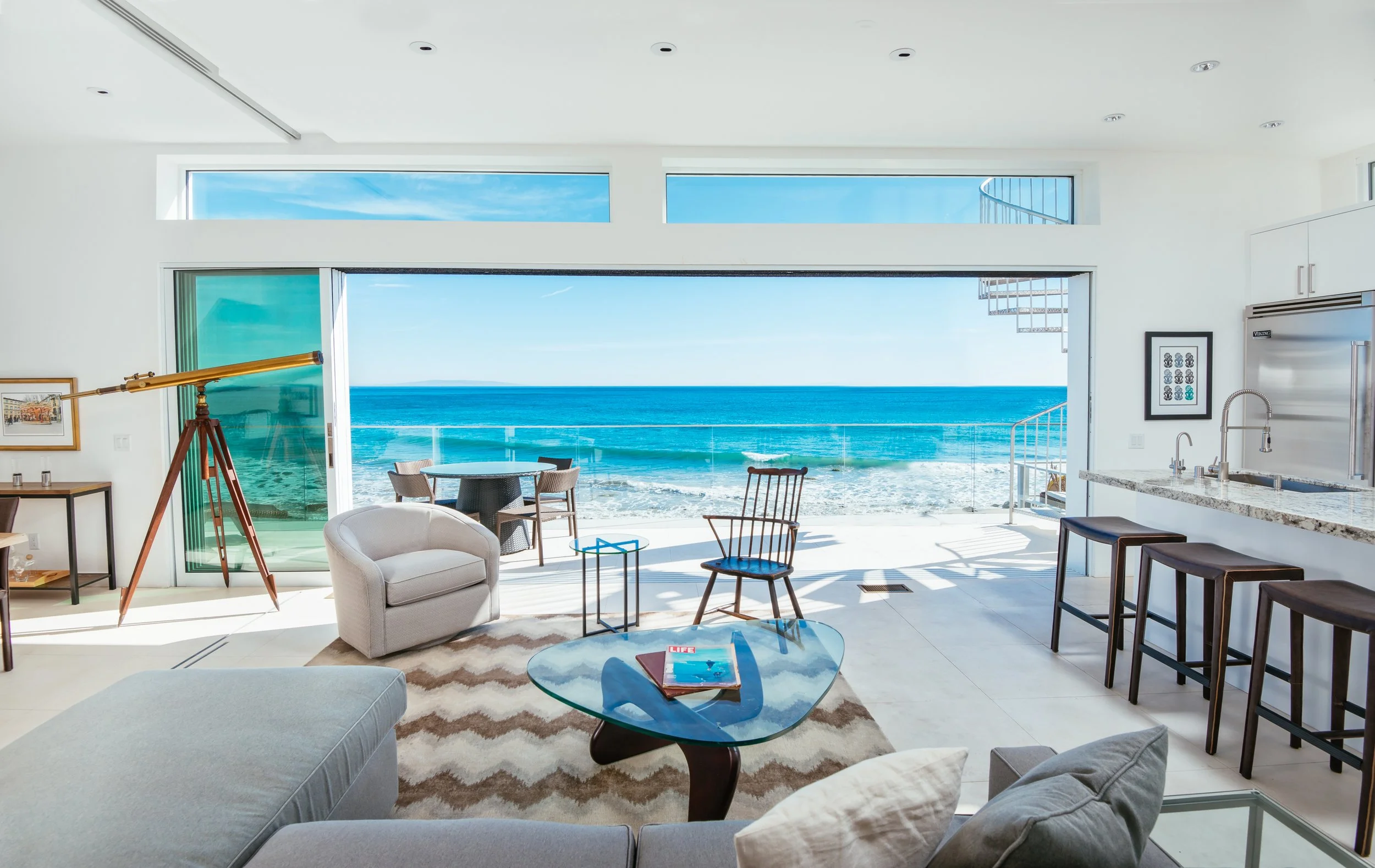Modern living room with large glass sliding doors opening to a beach view with blue sky, ocean waves, and a balcony with outdoor furniture.