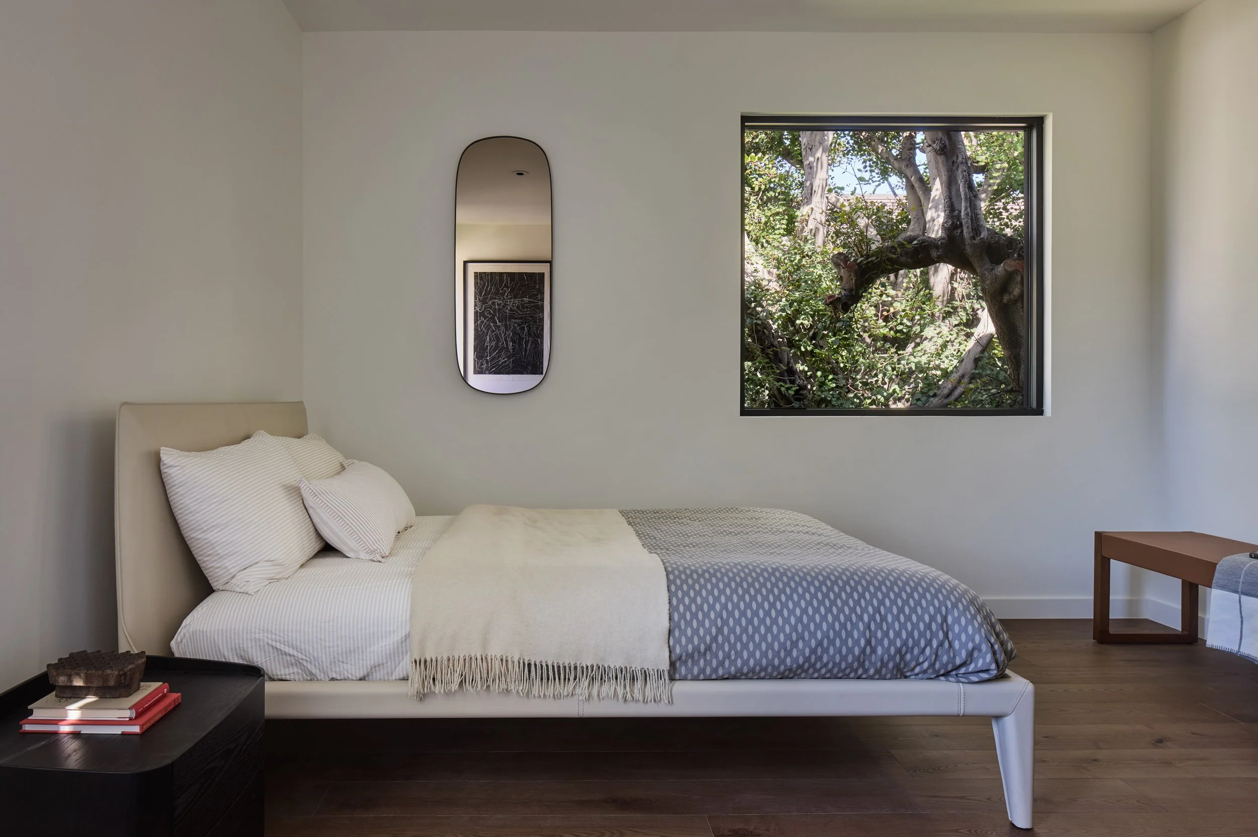A bedroom with a bed, white bedding, and pillows, a black nightstand with books and a decorative object, a large window with a tree outside, a mirror on the wall, and wooden flooring.