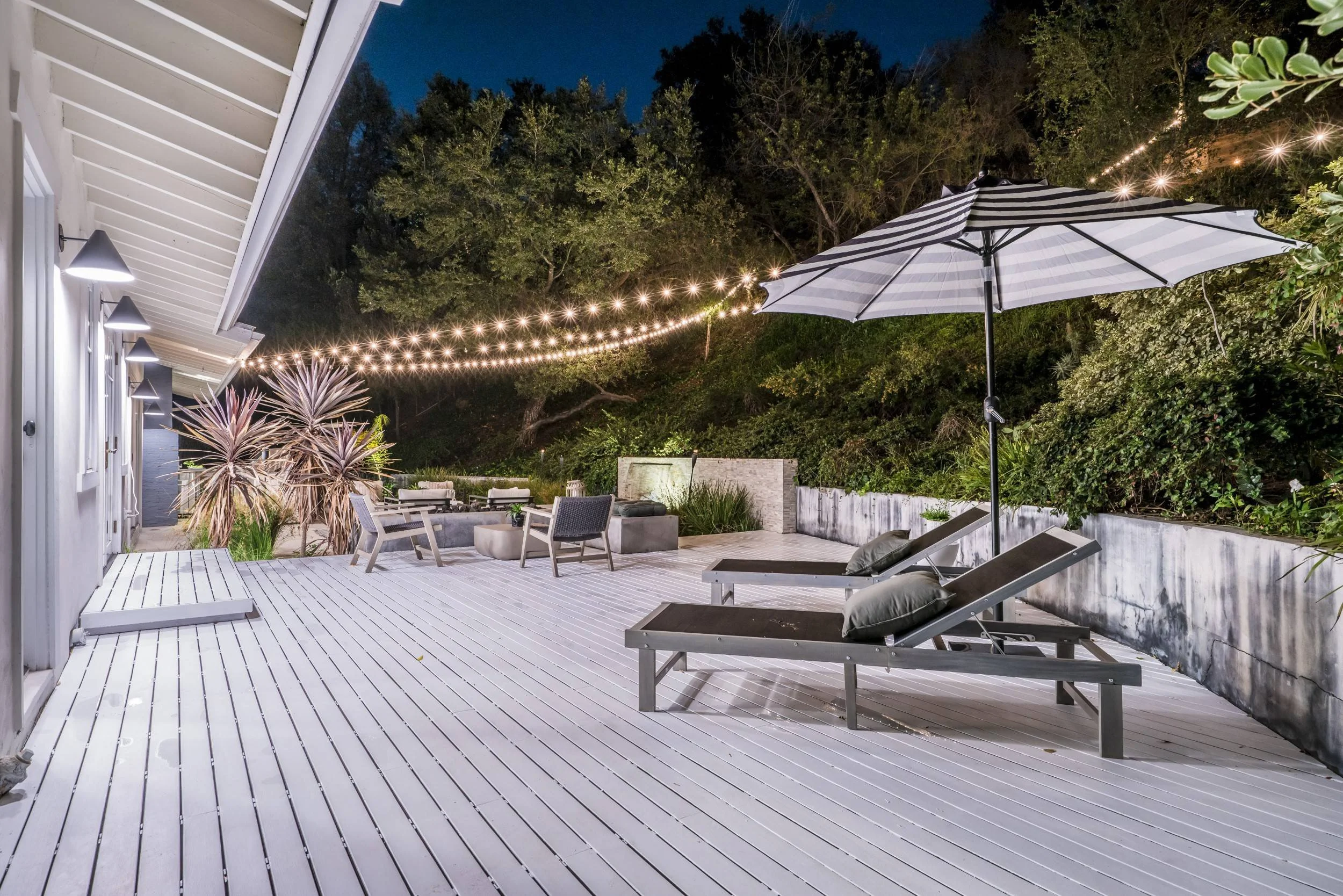 An outdoor patio with white wooden flooring, surrounded by greenery. There are lounge chairs, a large striped umbrella, a seating area with chairs and a firepit, string lights hanging above, and exterior wall lights on the house.