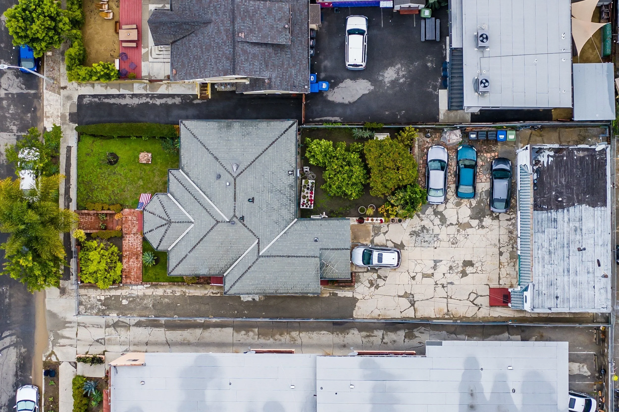 An aerial view of residential houses, parked cars, and a small yard with green grass and trees.