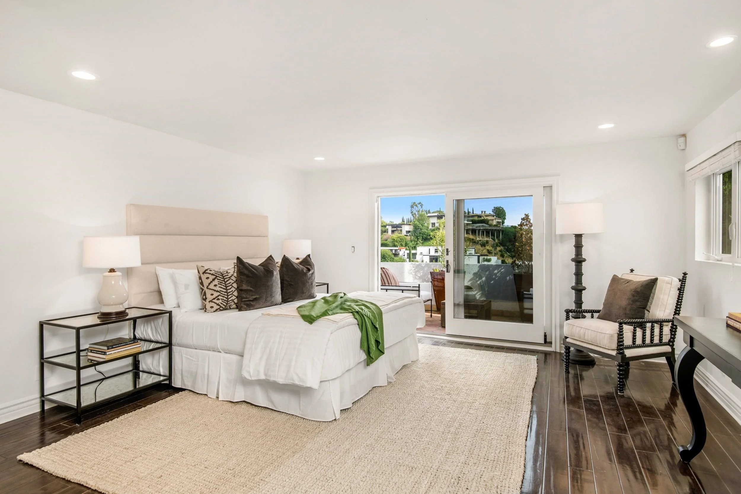 A bright living room with white walls and ceiling, featuring a white bed with pillows and a green blanket, a nightstand with books, a floor lamp, a black table, a seating chair, and a sliding glass door leading to a balcony with view of houses and tr