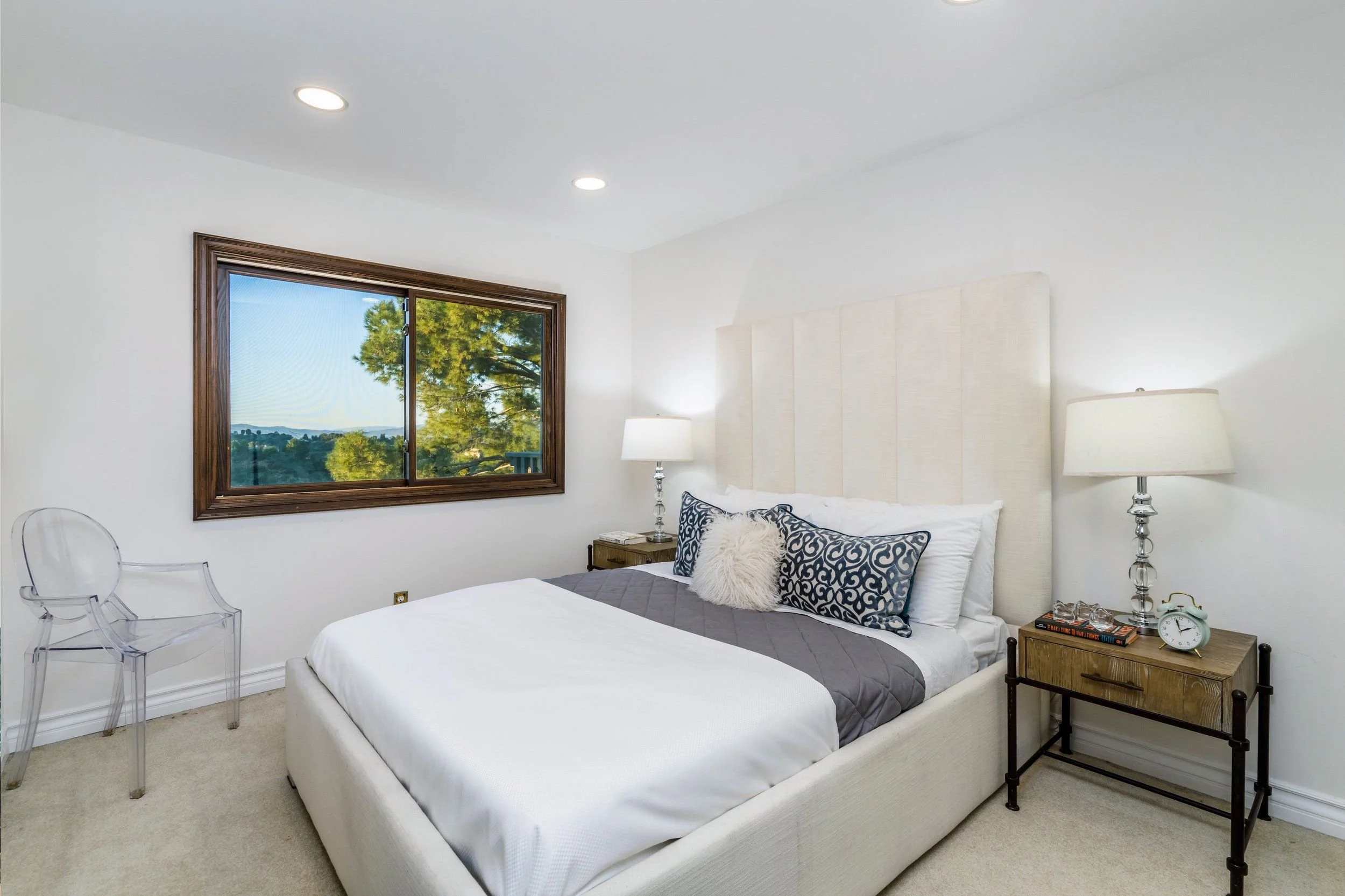 A bedroom with a white bed, decorative pillows, a nightstand with a lamp, books, a clock, and a window showing trees outside.