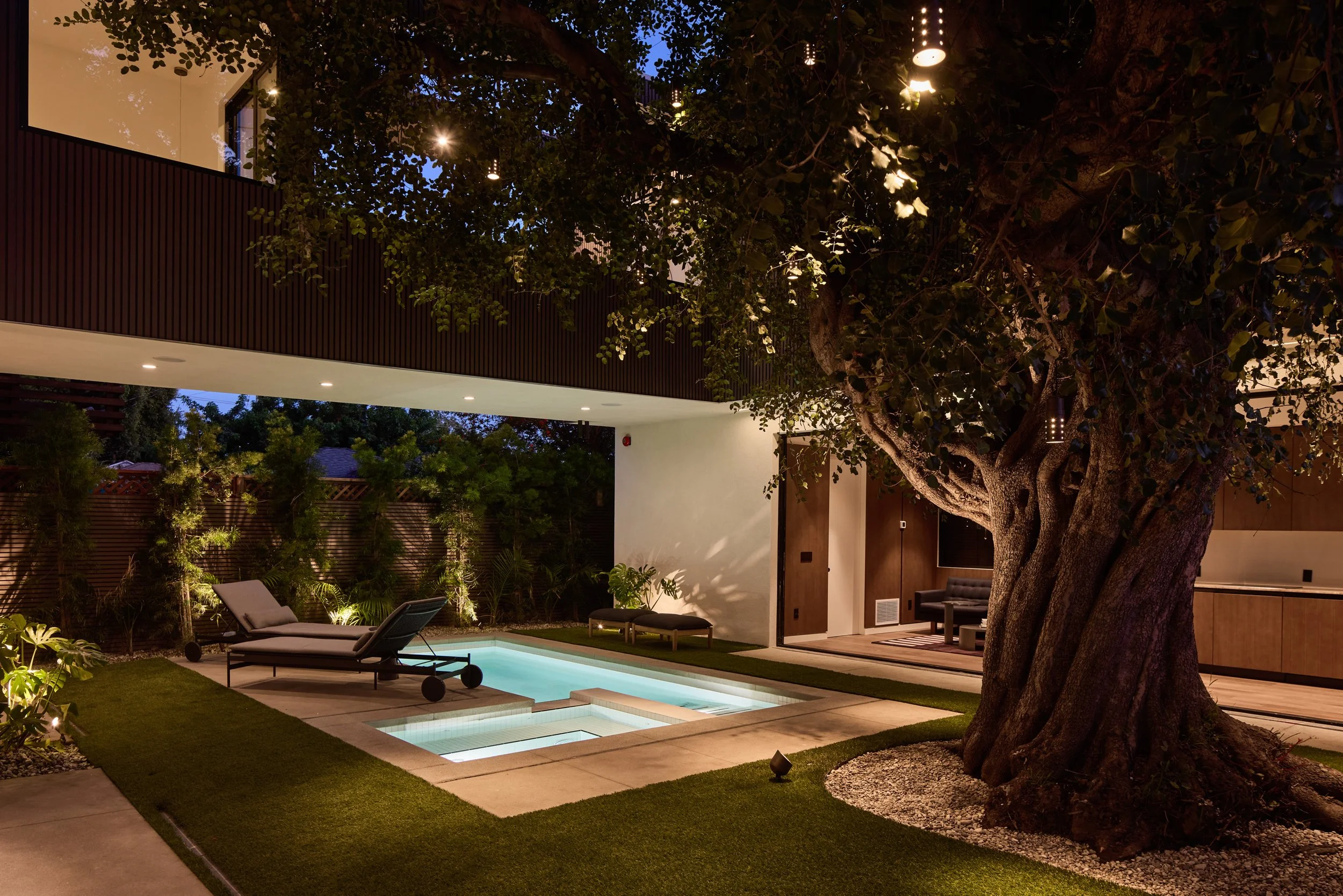A modern backyard patio at dusk with a large tree, outdoor seating, a small pool, and ambient lighting.
