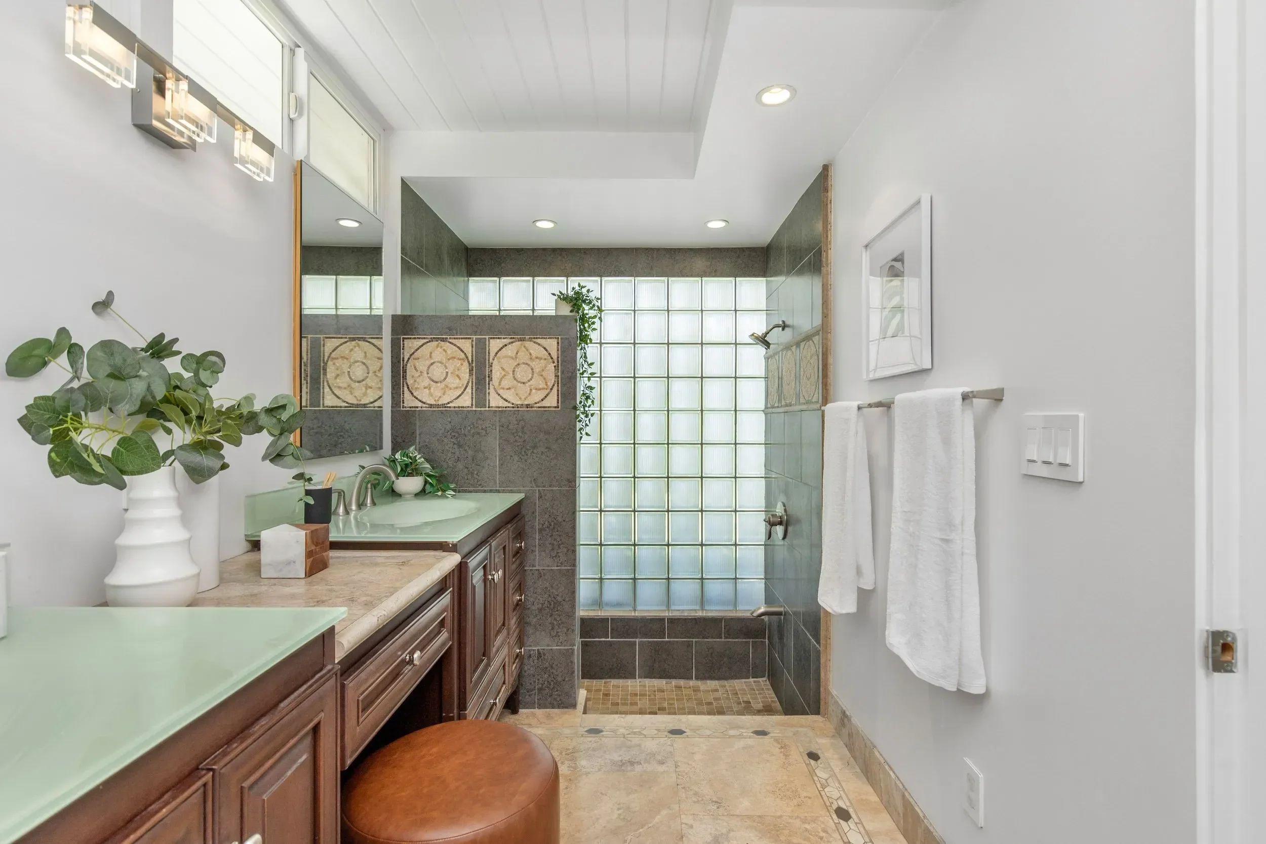 A bathroom with a vanity area that has a green countertop, wooden cabinets, and a mirror. To the left, there are decorative vases with green plants. In the background, there is a walk-in shower with a glass block wall and gray tiles, a small window, 