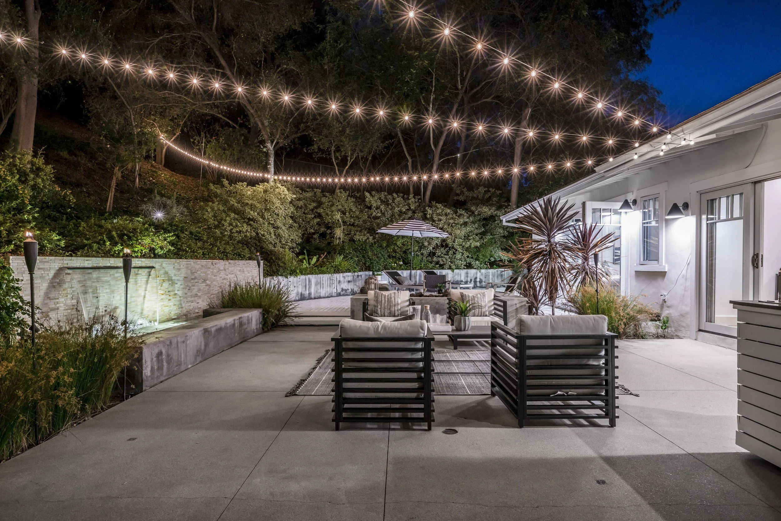 A backyard patio at night with outdoor string lights overhead, patio furniture including chairs and a table, a patio umbrella, surrounding greenery and trees, and a white house with windows and lights.