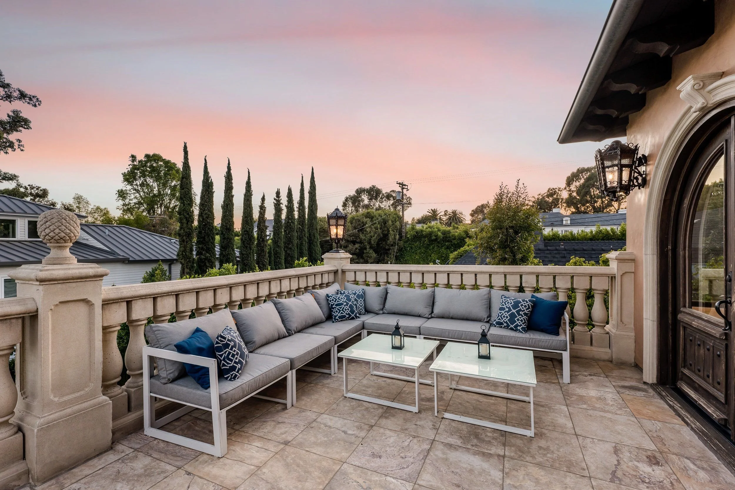 An outdoor rooftop patio with a sectional sofa with pillows and a coffee table, surrounded by a decorative beige railing, with trees and a pastel pink and purple sky in the background.