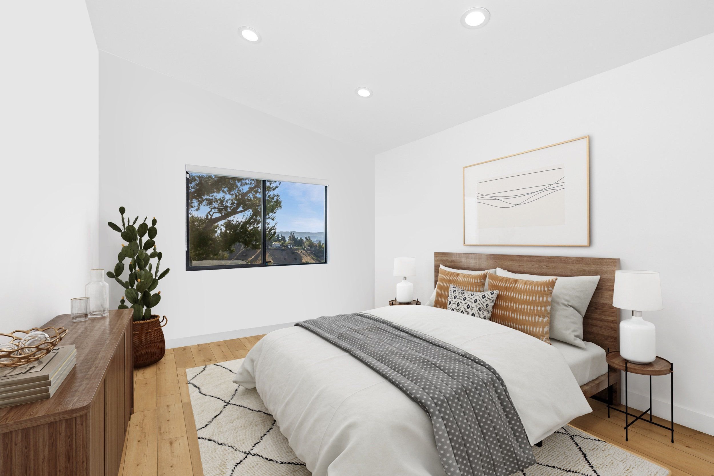 A bedroom with white walls, a large window showing trees and sky outside, a wooden bed with white bedding and decorative pillows, two white bedside lamps, a wooden dresser with glass vases and glasses, a framed abstract artwork, a black and white rug