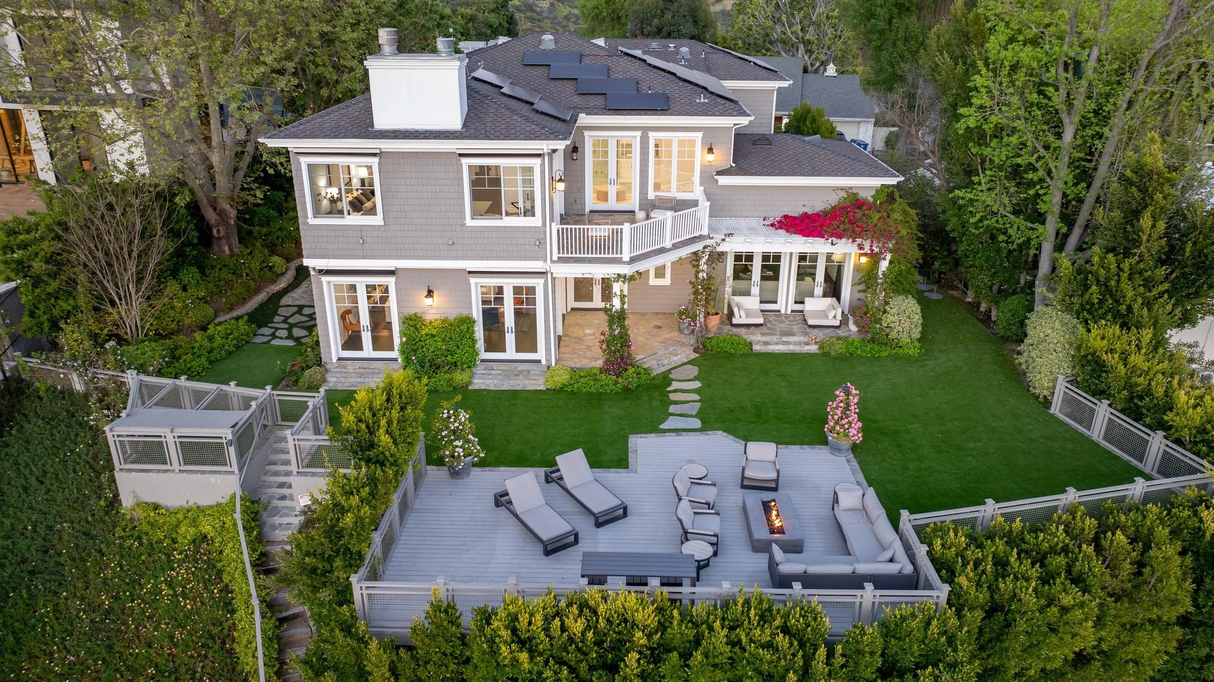 Aerial view of a large two-story house with a backyard. The yard features a wooden deck with lounge chairs, a fire pit, and outdoor seating. The house has multiple windows, a small balcony, and solar panels on the roof. Surrounding the yard are trees
