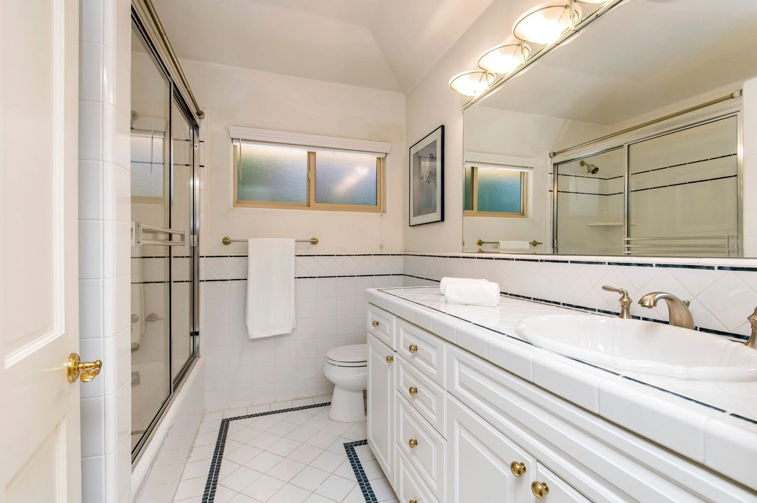 A bathroom with a white vanity, gold knobs, a double sink, and a large mirror. There are frosted windows, a shower with glass doors, and a toilet. The floor and walls are tiled with white tiles accented by black border tiles.