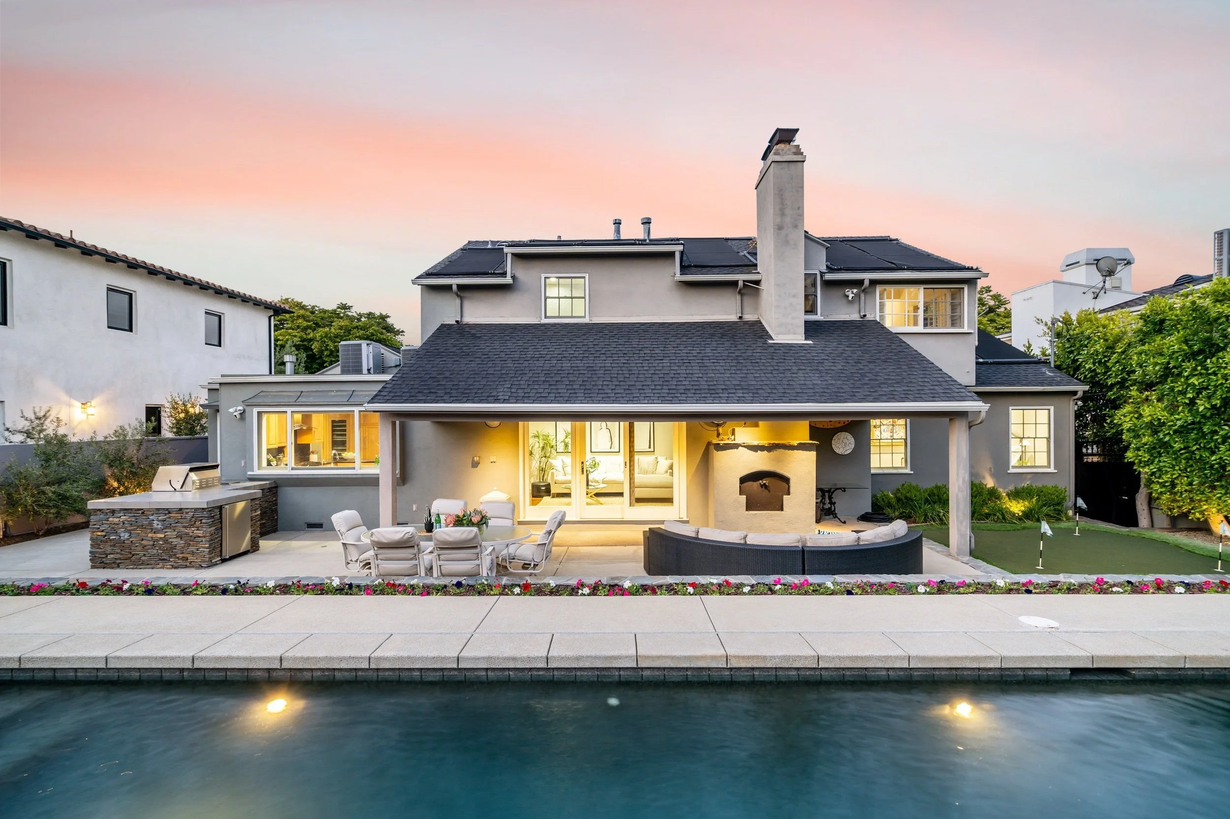 Backyard of a modern house at sunset, overlooking a swimming pool with lights, outdoor furniture including a table, chairs, and a curved lounge, surrounded by a stone fire pit and flower bed, with neighboring houses and trees in the background.