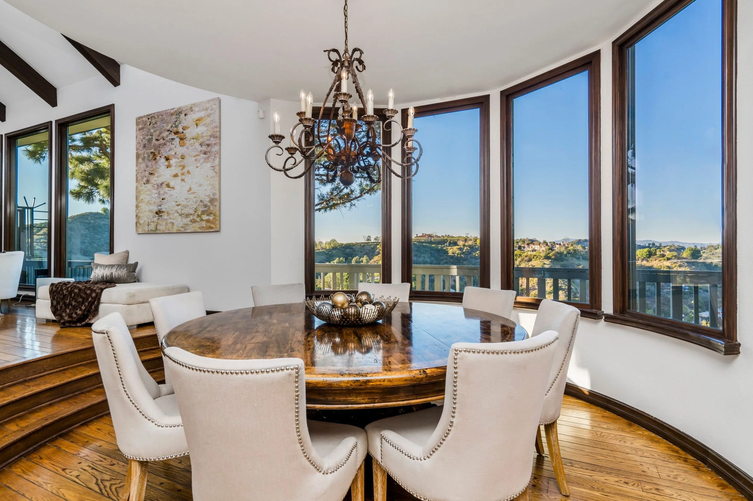 A dining room with a round wooden table surrounded by white upholstered chairs, a chandelier hanging from the ceiling, large windows showing a scenic view of greenery and hills, and a partial view of a living area with a white sofa and abstract paint