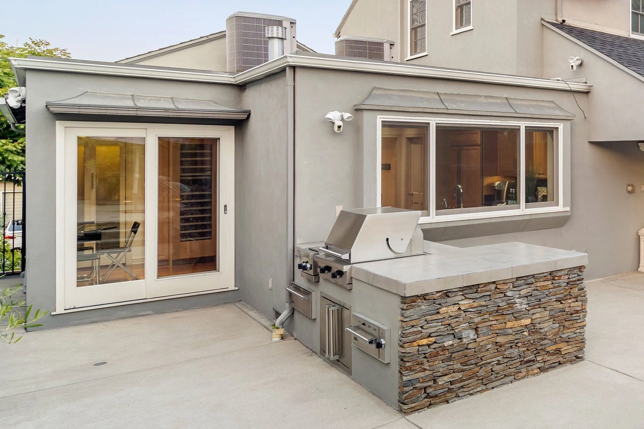 Backyard patio area with a built-in outdoor kitchen featuring a grill, stone accents, and a sliding glass door leading into the house. There are two wall-mounted security cameras and a window on the house's exterior.