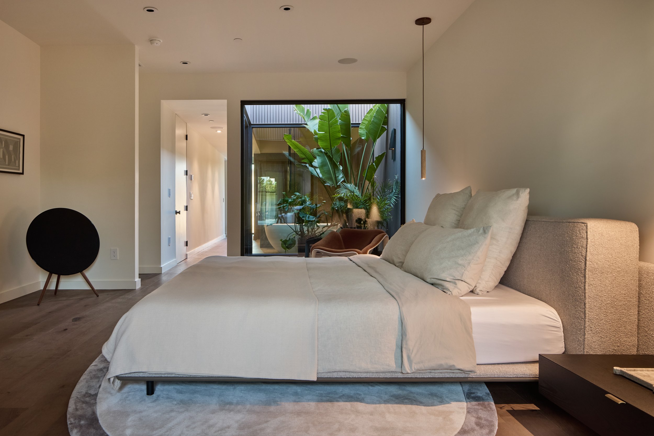 A modern bedroom with a large bed with beige bedding, a sitting area with a plant in front of a large glass door, and minimal decorating.