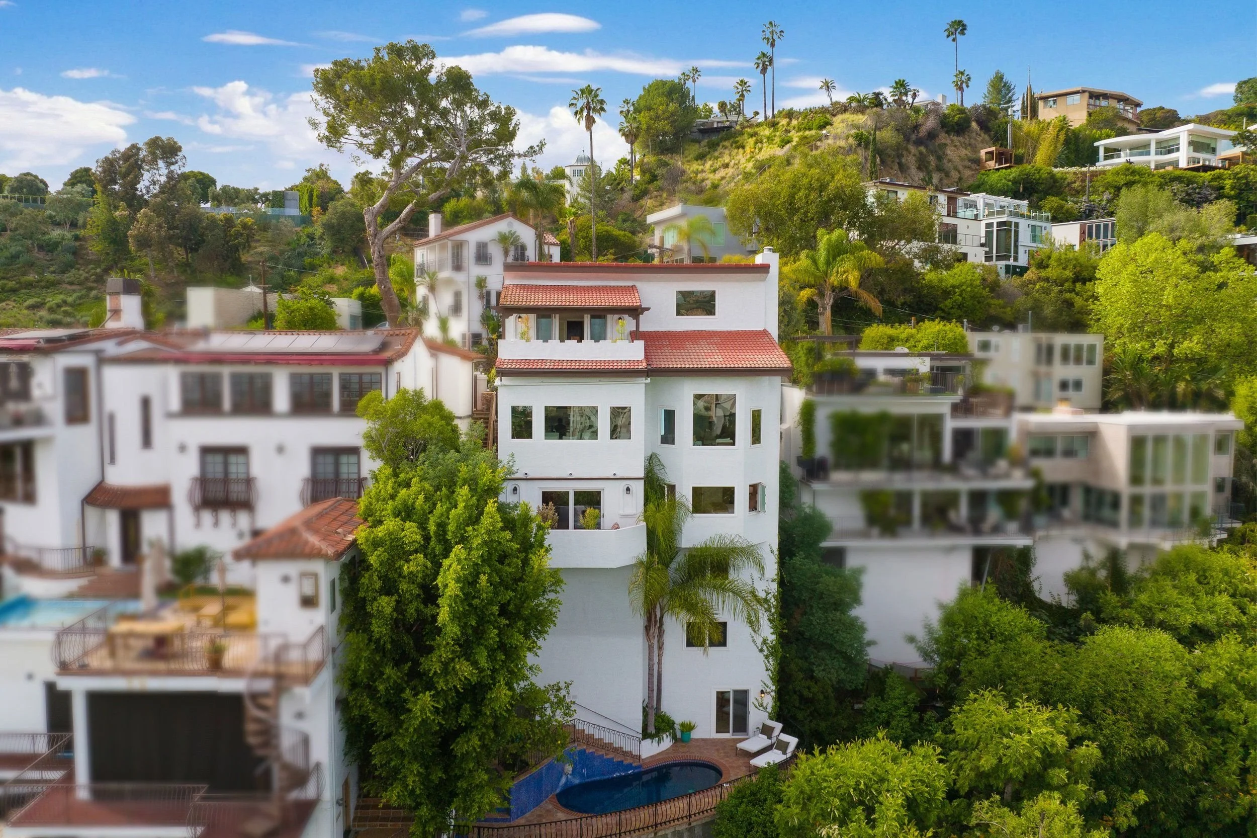Residential hillside neighborhood with white houses, lush green trees, and tall palm trees under a partly cloudy blue sky.