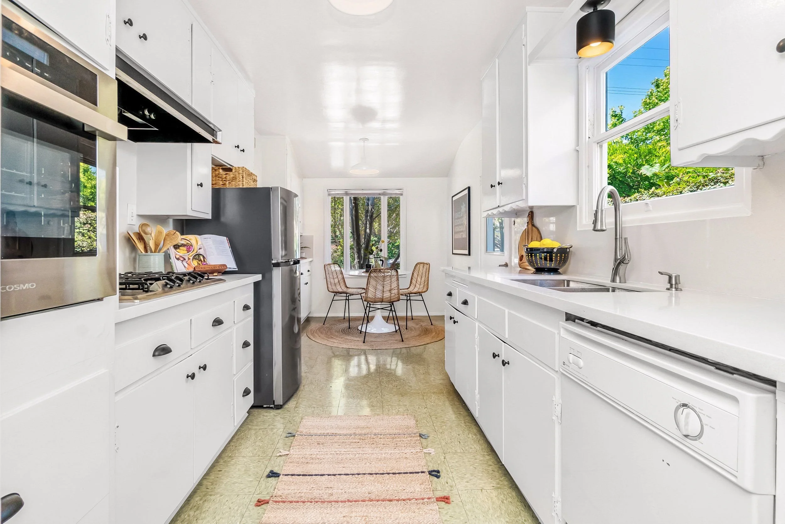 White kitchen with black cabinet handles, stainless steel refrigerator, window with greenery outside, dining area with woven chairs, small round table, and kitchen countertop with a black bowl of lemons.