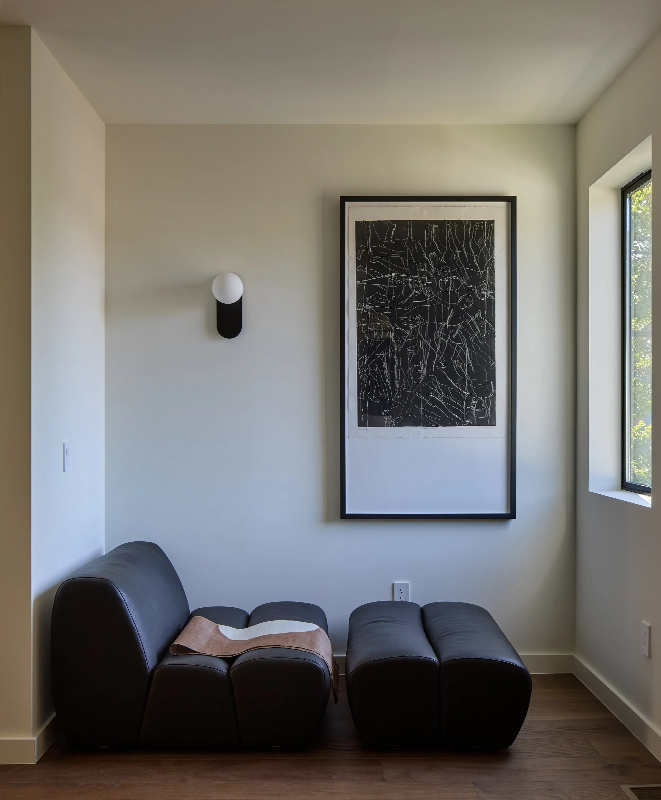 A black leather sectional sofa with a tan throw blanket on one end, located in a modern living room. The wall above features a large, framed black artwork with white abstract lines, and a black and white wall sconce light is mounted on the wall. Ther