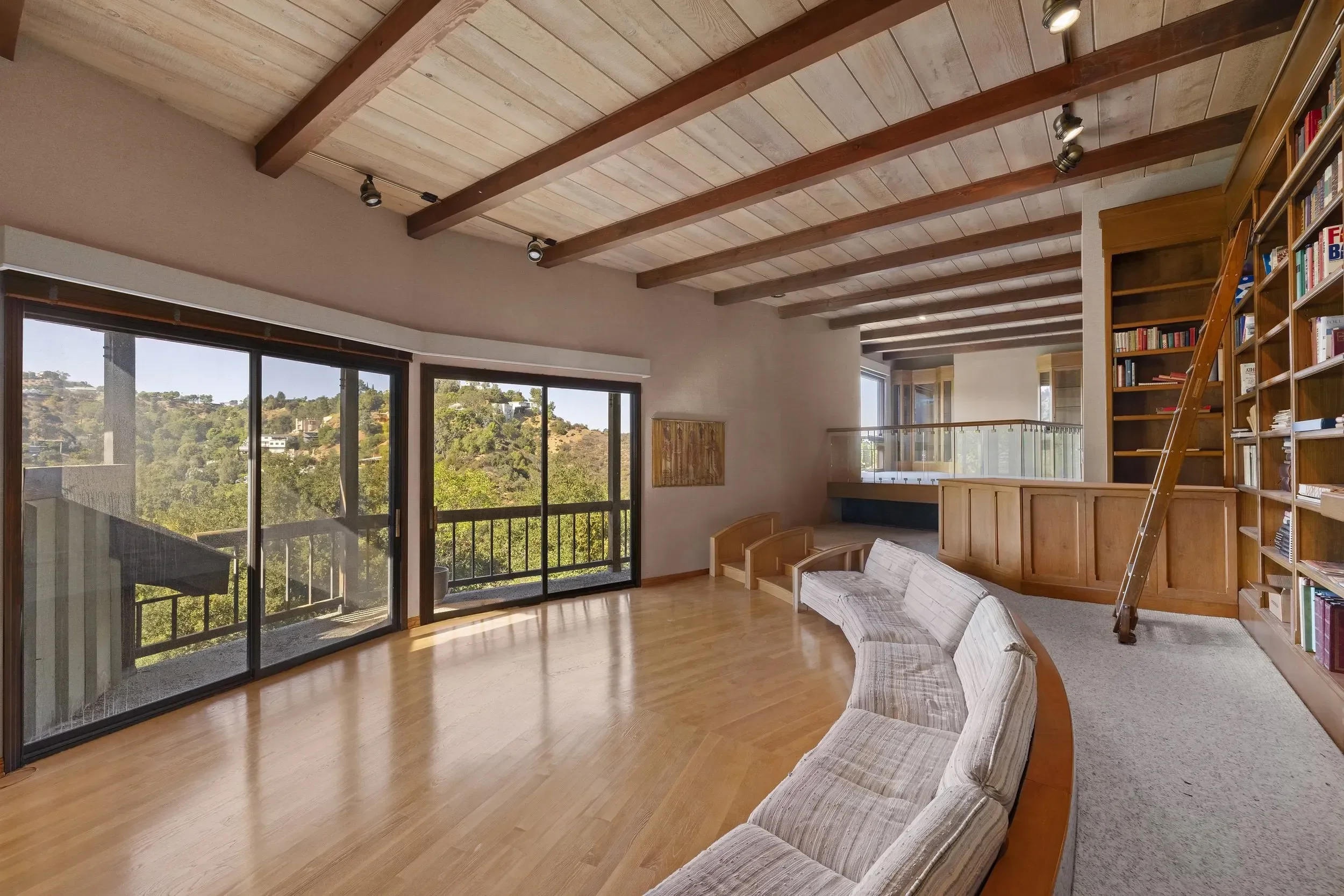 Living room with large windows, wooden beams ceiling, bookshelf, ladder, and a couch.