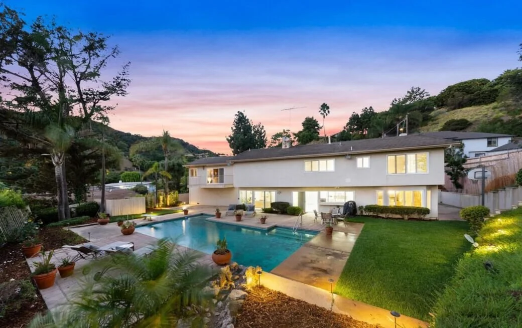 Backyard view of a two-story house with illuminated windows, a swimming pool, and outdoor seating, surrounded by lush green landscaping during sunset.
