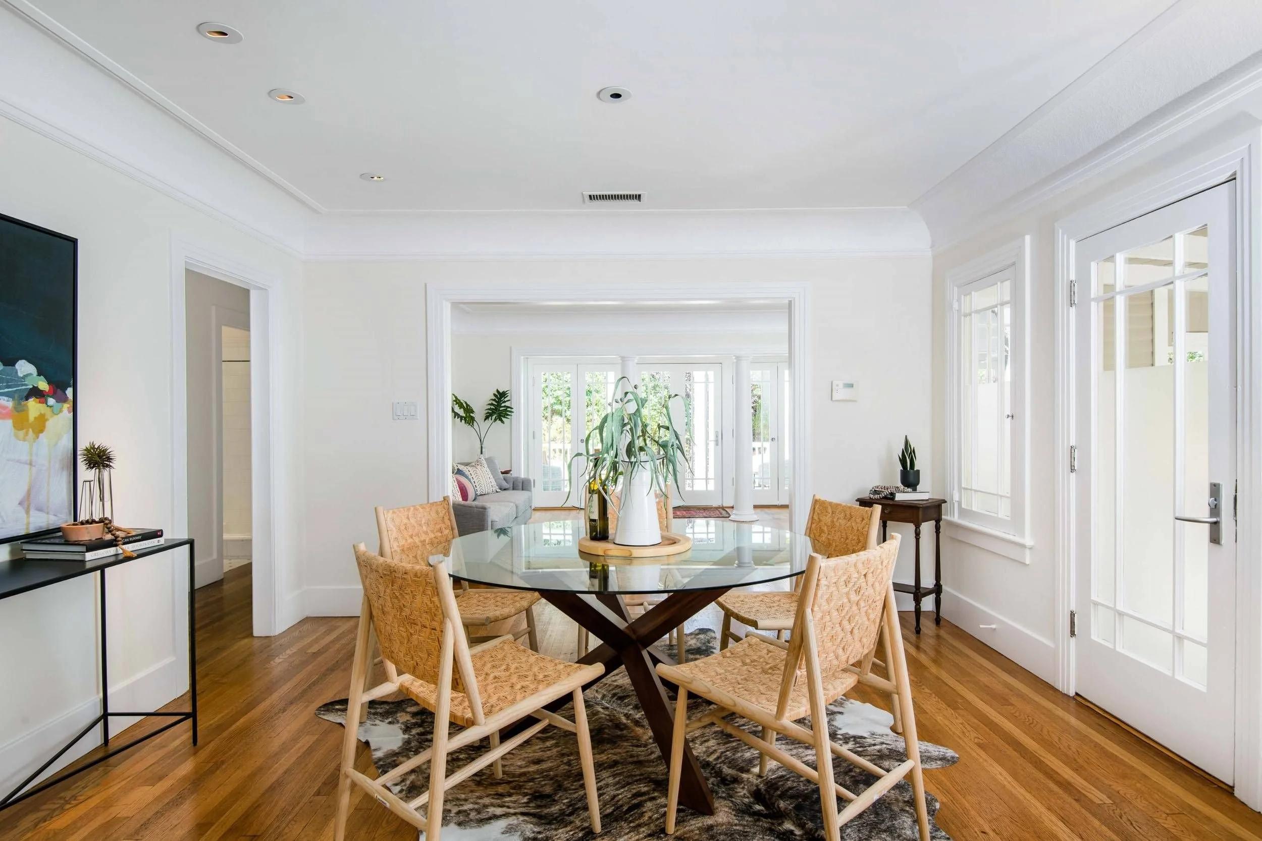 A dining area with a round glass table surrounded by six woven rattan chairs. Toward the back, a living room with a gray sofa, pillows, and potted plants can be seen through an open doorway. The room has white walls, wooden flooring, multiple windows