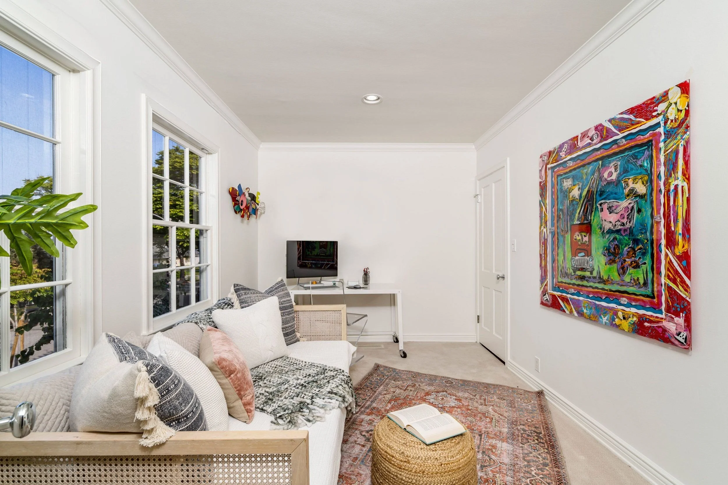 Bright living room with white walls, a colorful large abstract painting, a white sofa with patterned cushions, a rattan ottoman with an open book, a small white desk with a computer, and multiple windows showing trees outside.
